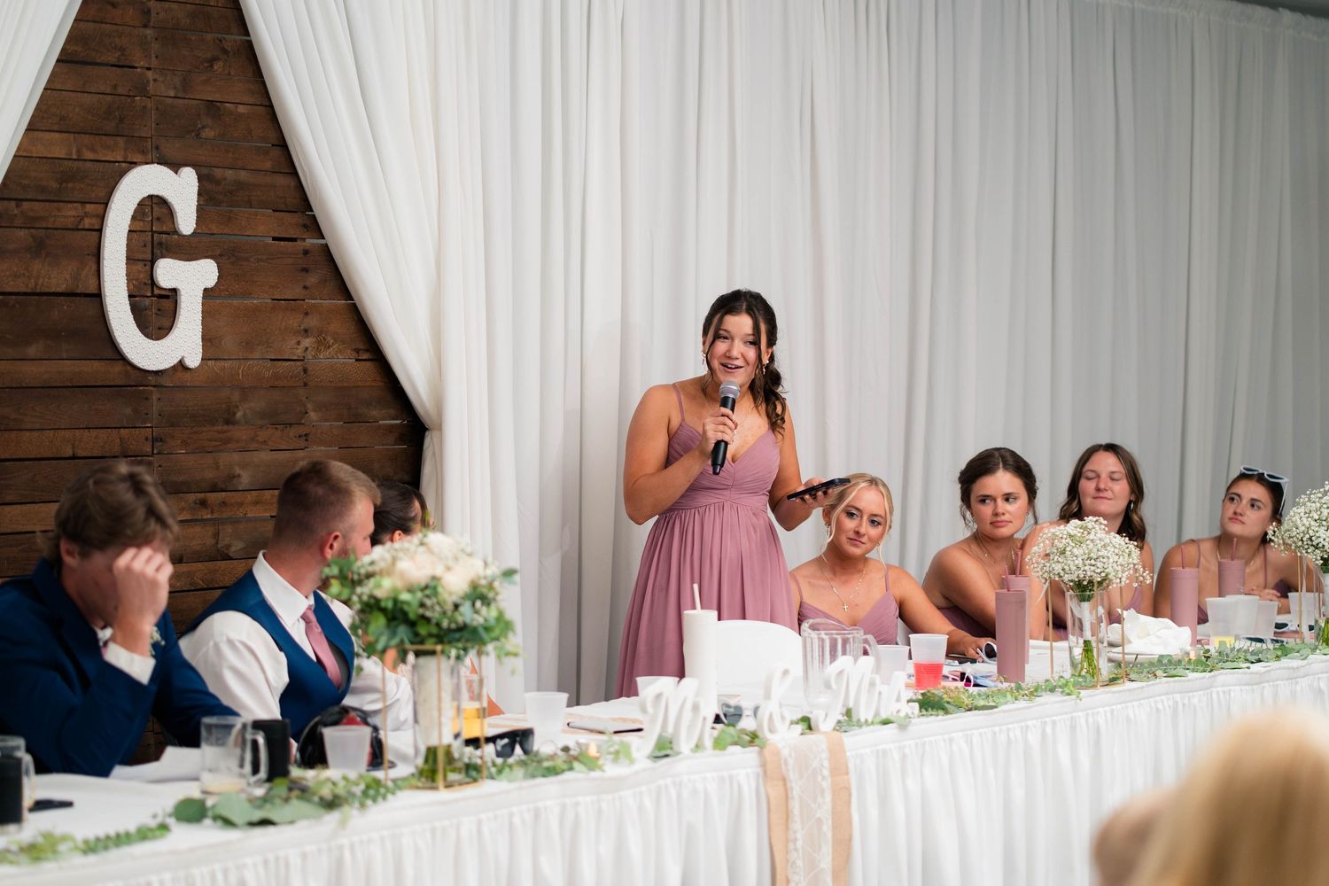Bridesmaid in mauve dress delivers heartfelt speech at wedding reception while bridal party listens and smiles.