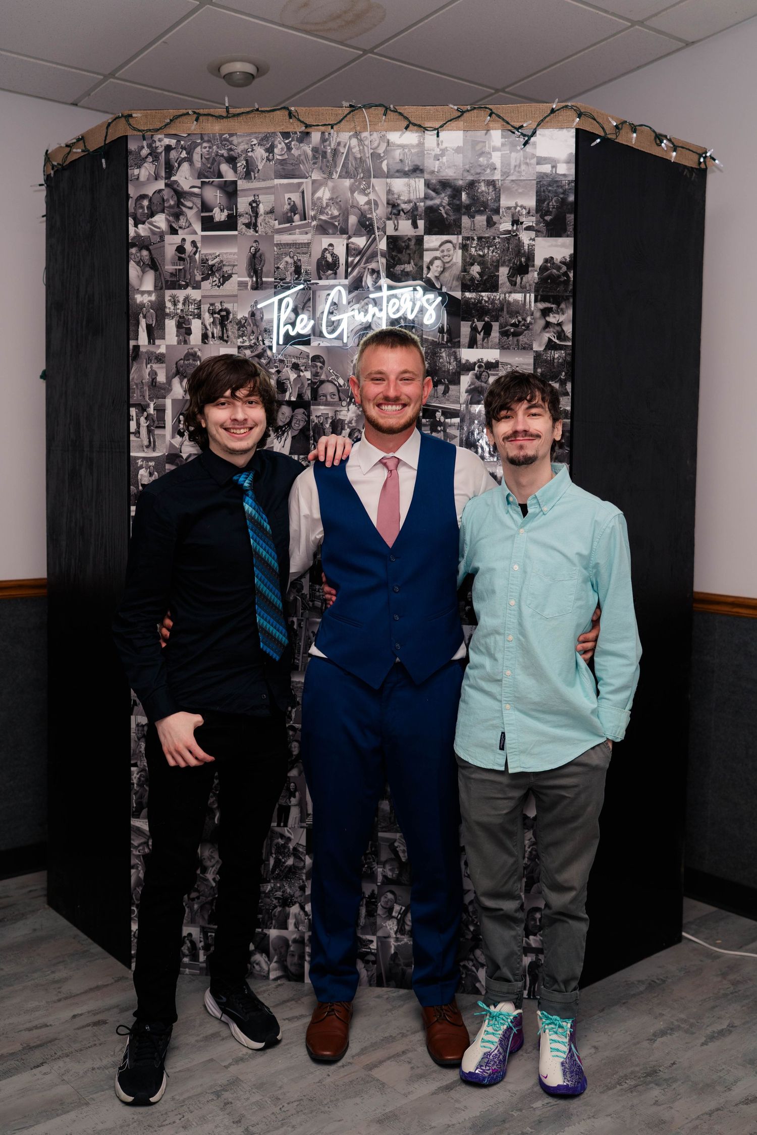 Three men pose smiling in front of a wedding photo collage backdrop with a neon sign reading The Garters.