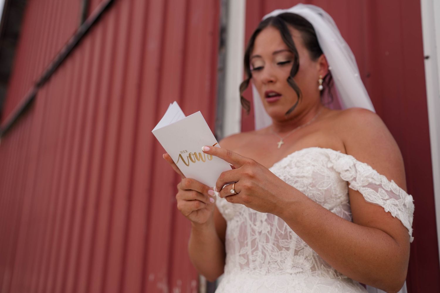 Bride in lace off-shoulder gown and veil reading a heartfelt letter outside a red barn.