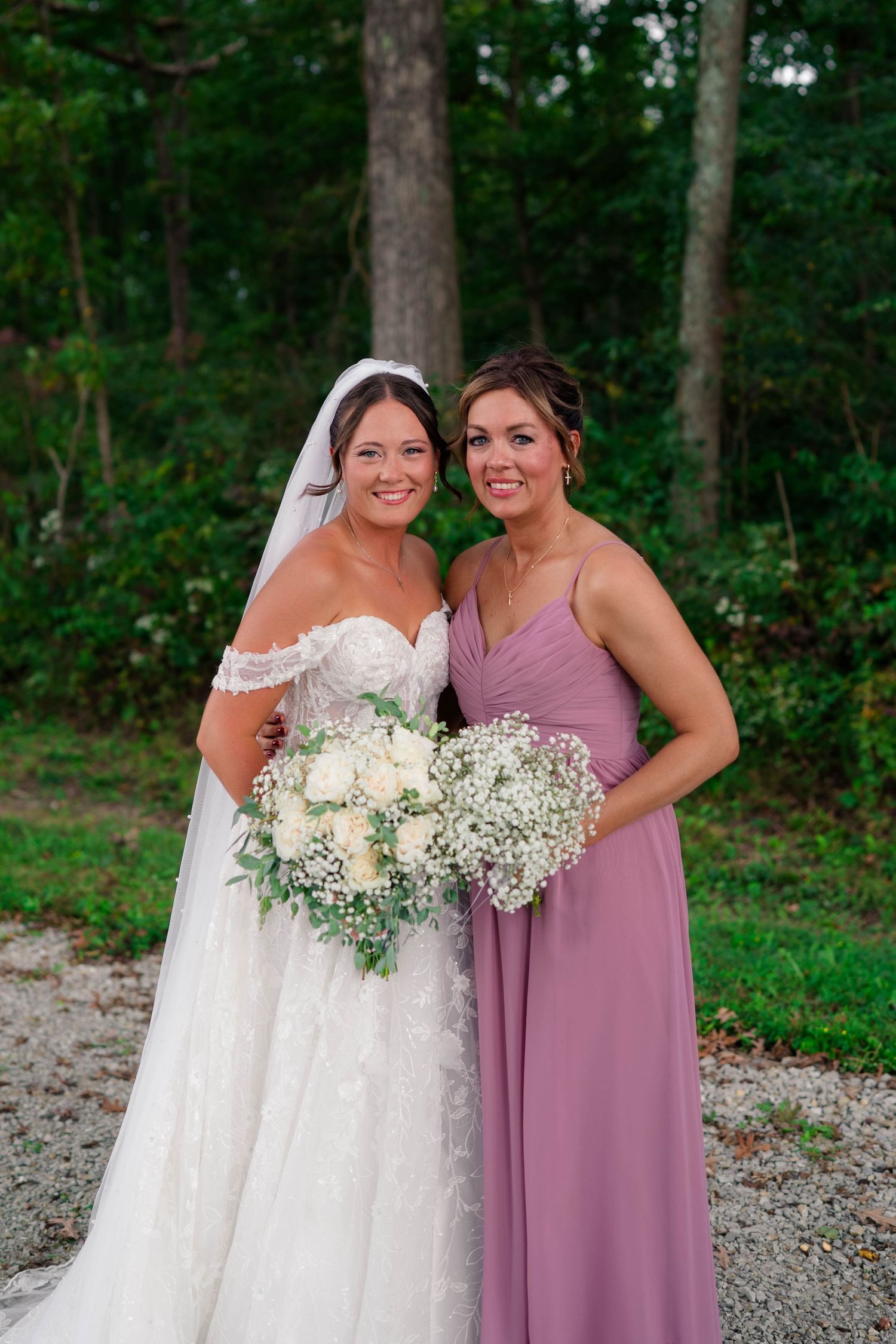Smiling bride in lace off-shoulder gown stands with bridesmaid in dusty rose dress holding floral bouquets.