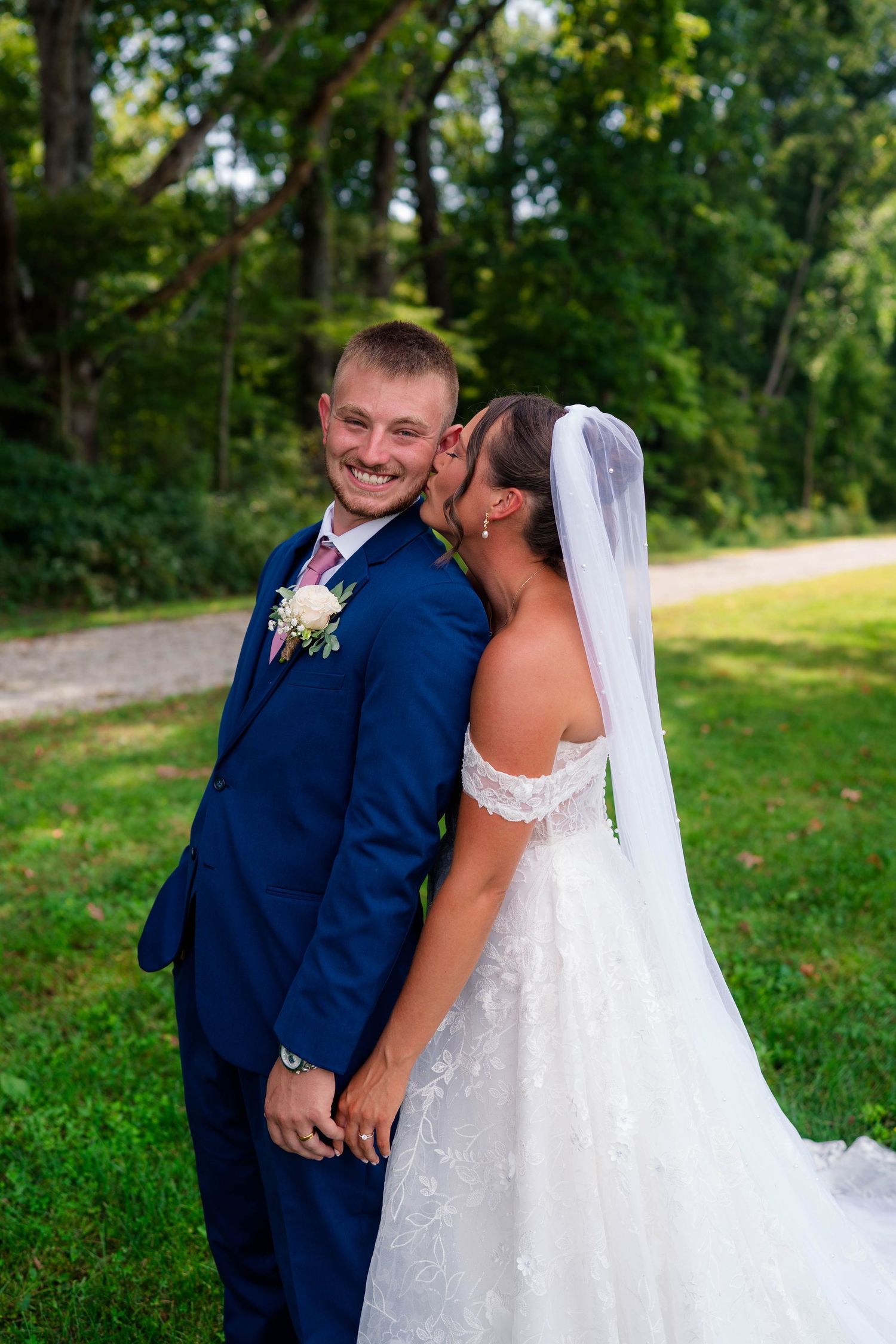 Bride kissing groom on cheek as he smiles in navy blue suit with boutonniere outdoors.