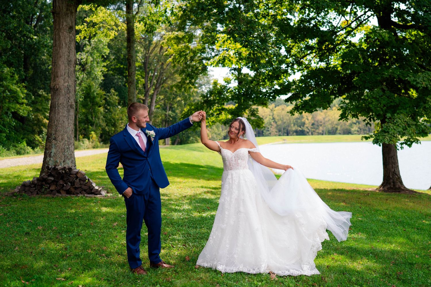 Groom twirling bride in white lace gown on green lawn near a lake under a large tree.