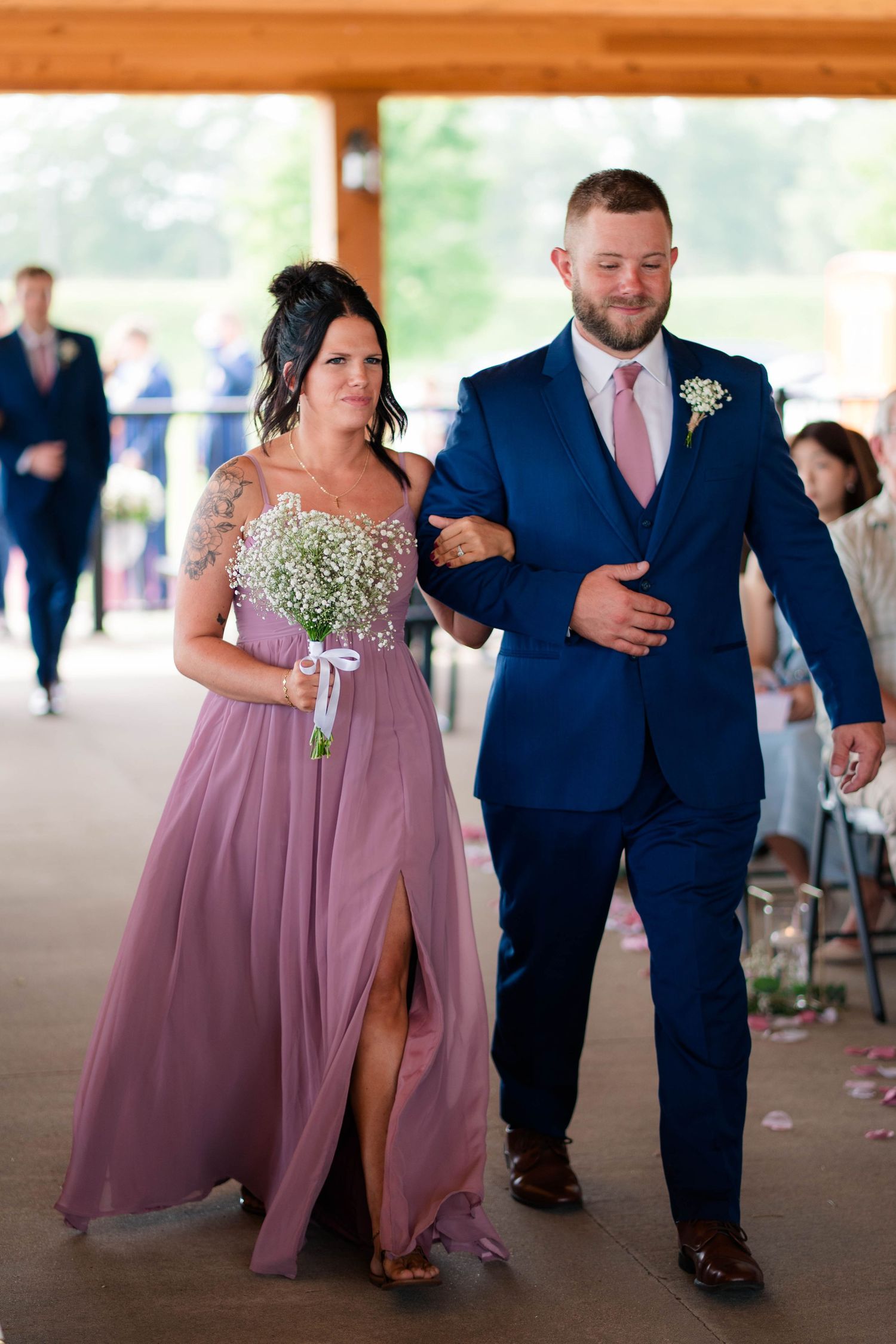 Groomsman in navy suit escorts bridesmaid in mauve gown down wedding aisle.