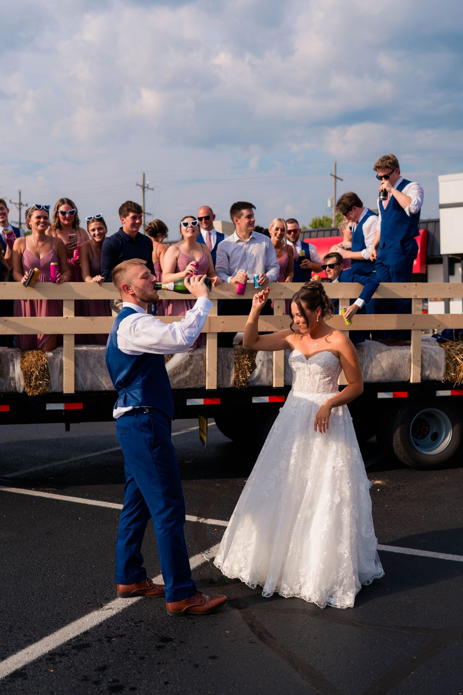 Groom spins bride in white strapless gown while wedding party celebrates on flatbed truck behind them.