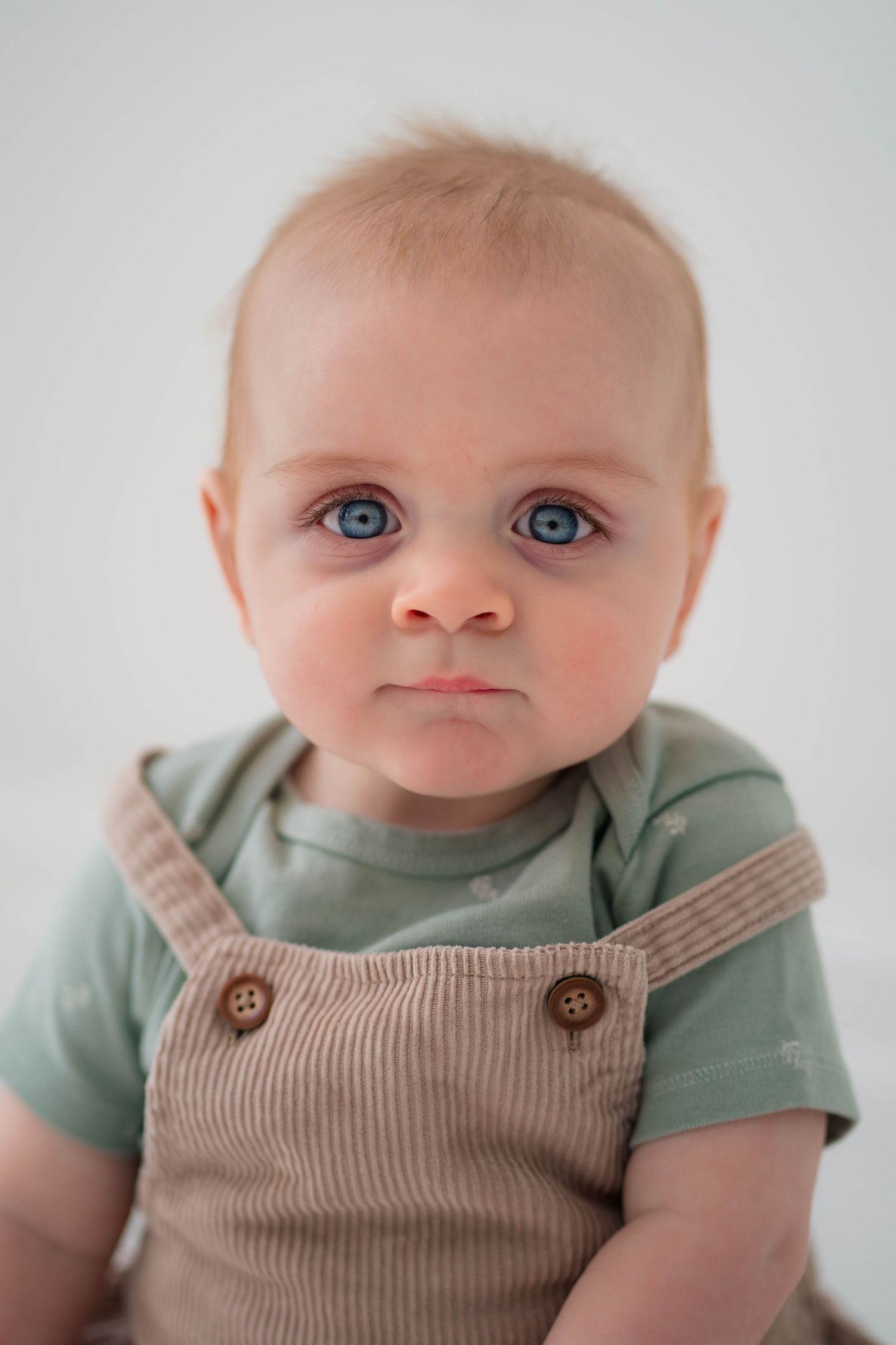 Adorable baby with blue eyes and light hair wearing a green onesie and brown corduroy overalls against white background.