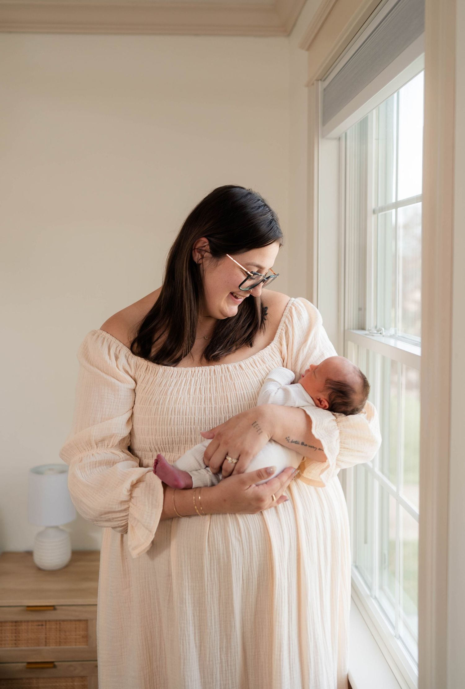 Mother in cream off-shoulder dress tenderly gazes at her newborn baby by a bright window.