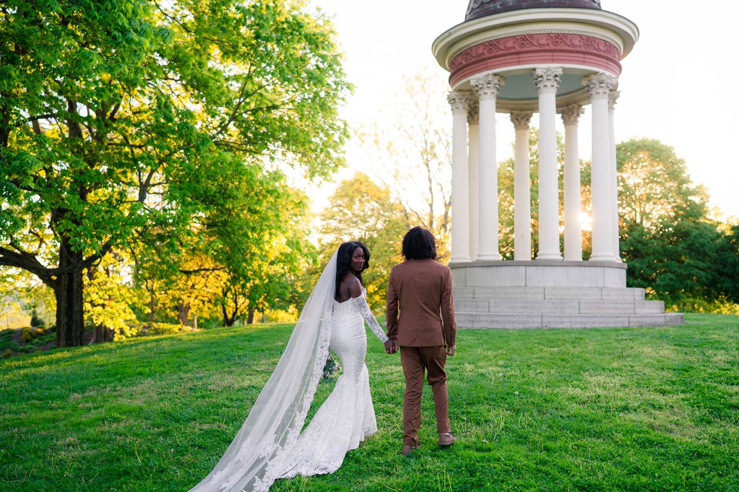 Bride in white gown and groom in brown suit stand before a classical rotunda in a lush green park at sunset.