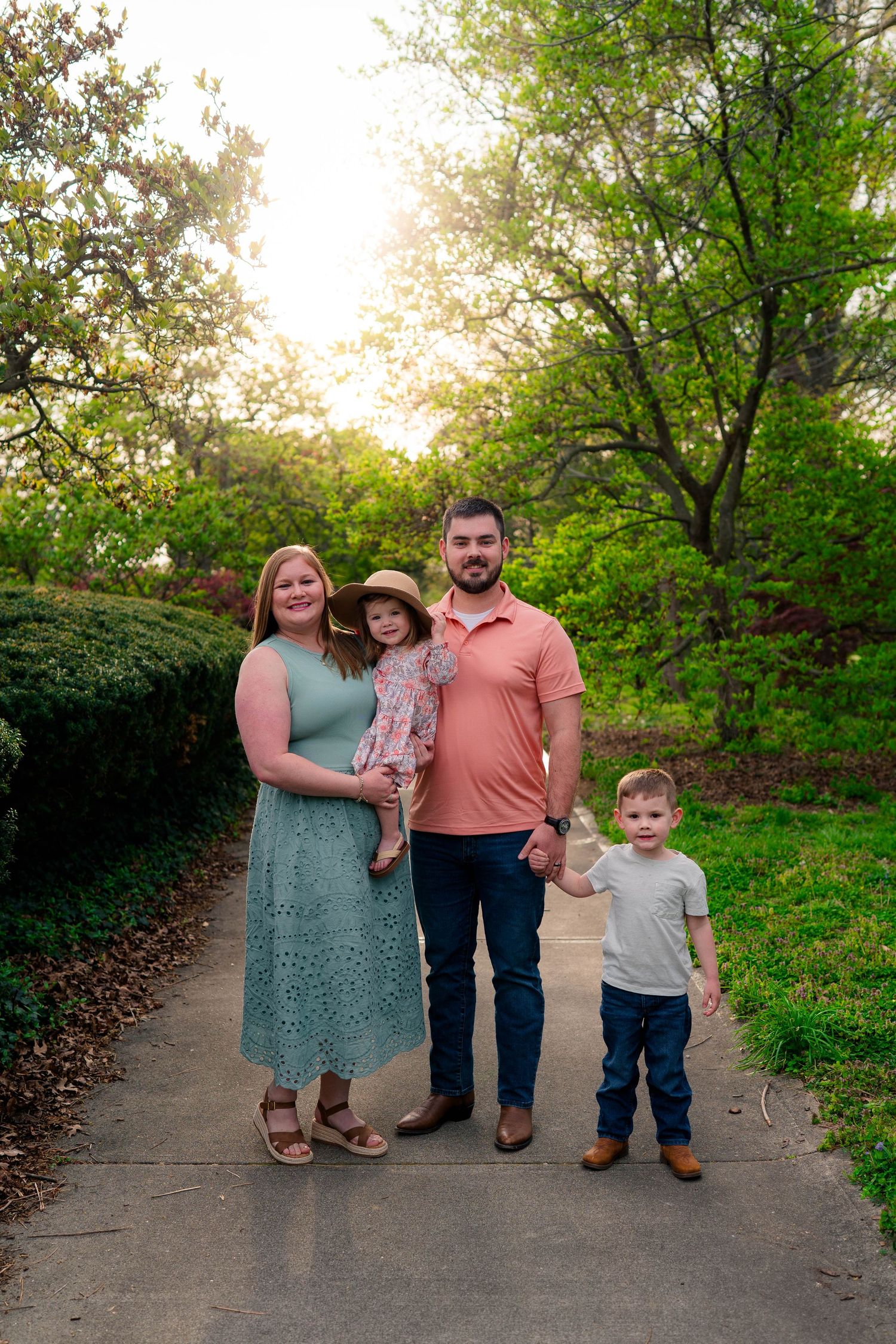 A family of four stands on a sunlit garden path, mom in teal dress, dad in pink shirt, with two young children.