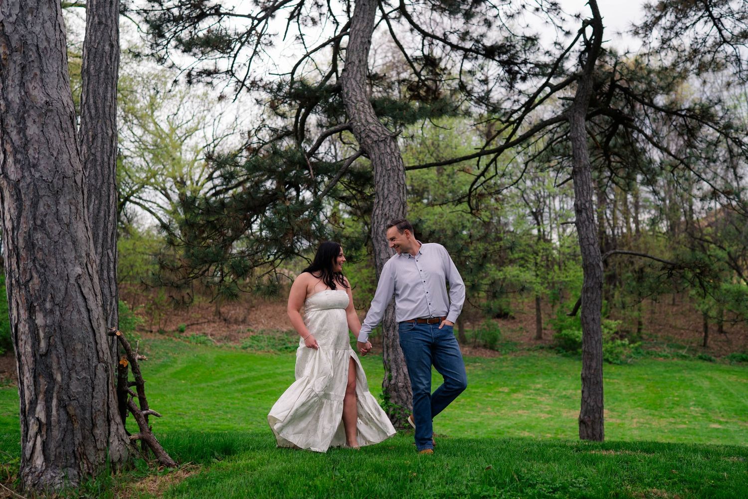 Couple holding hands walking through lush green park among tall pine trees during spring engagement photo session.