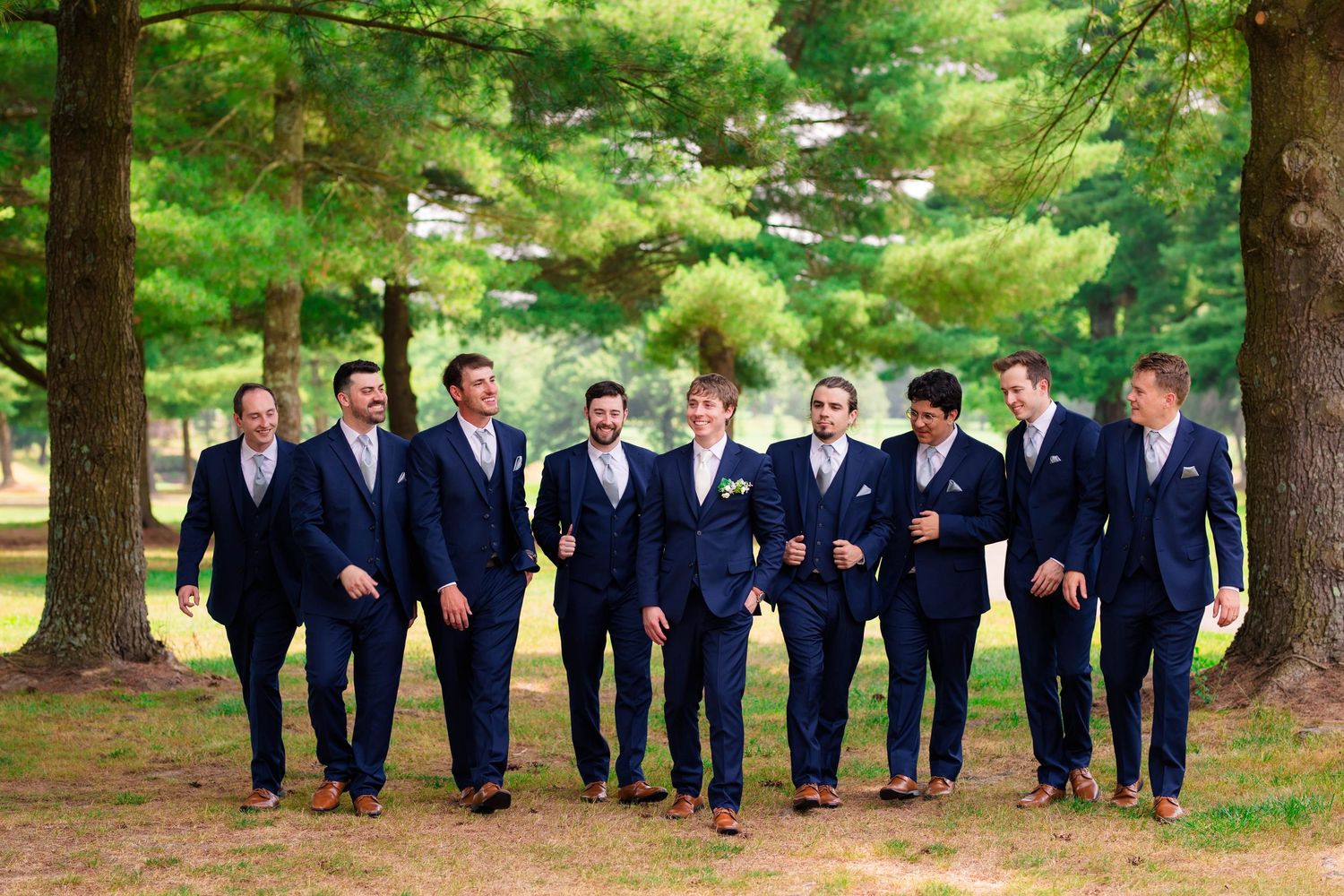 Groom and groomsmen walking confidently in navy suits through a tree-lined path on a sunny wedding day.
