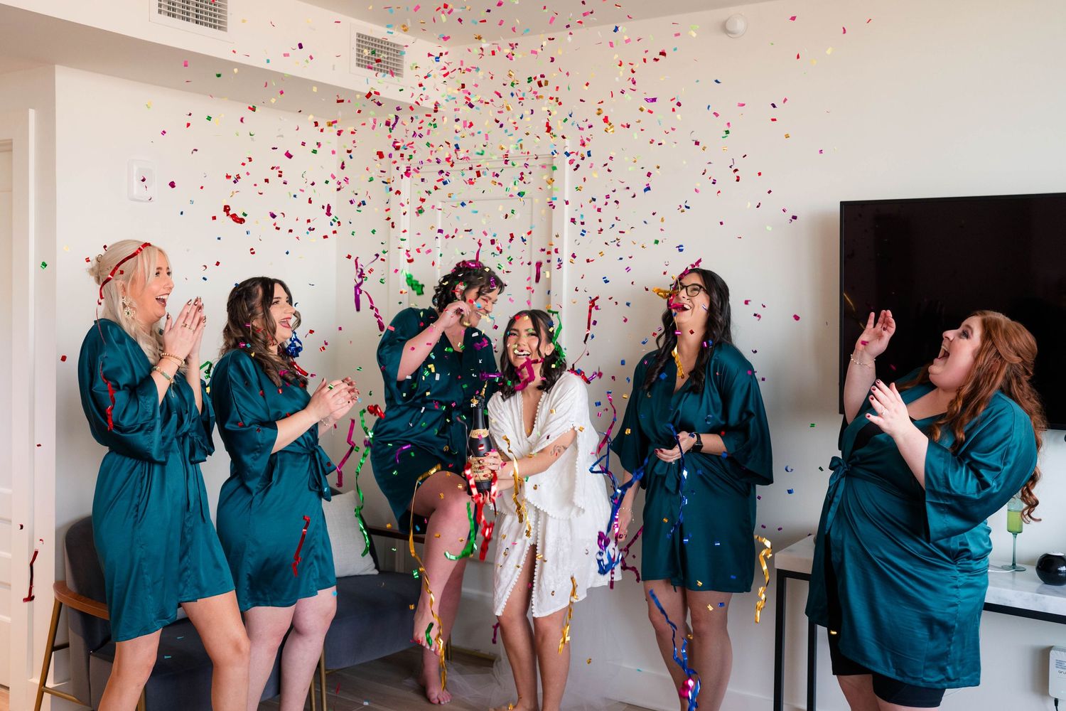 Bride and bridesmaids in matching teal robes celebrate with confetti cannon during wedding morning preparations.