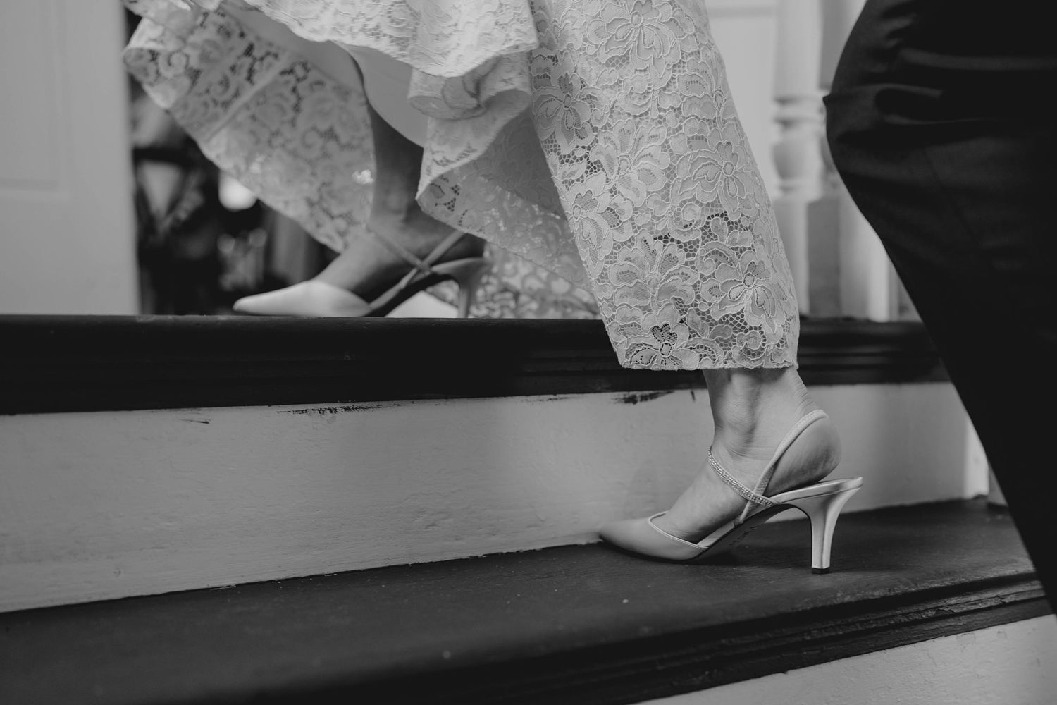 Black and white close-up of bride wearing white heels on dark wooden staircase at wedding.