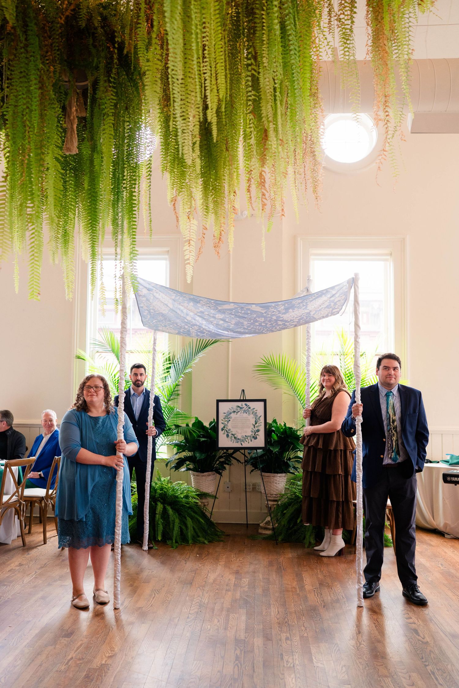 Wedding party stands beneath a chuppah decorated with hanging ferns and tropical greenery indoors.