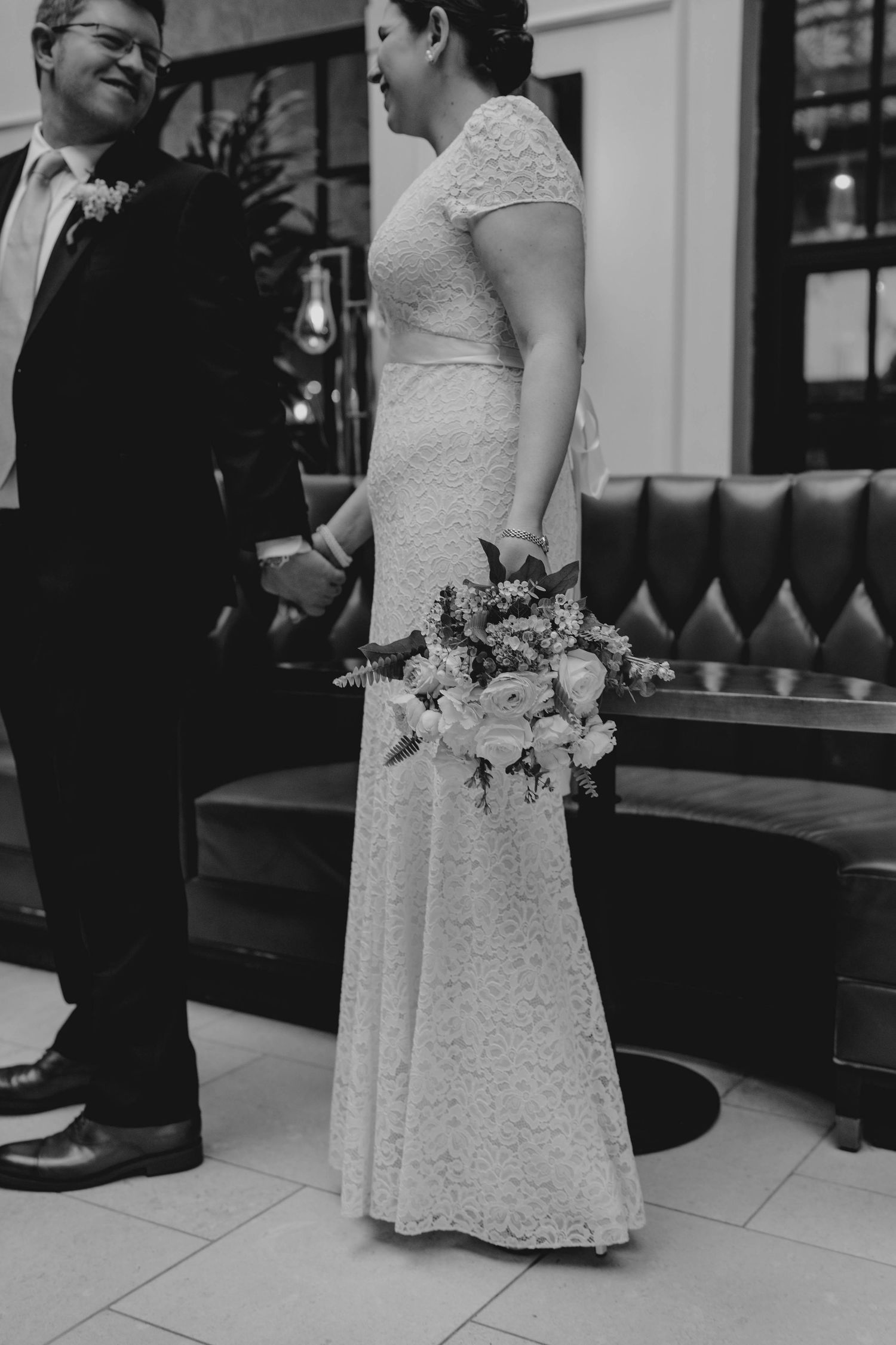 Black and white photo of bride holding bouquet and groom holding hands at wedding venue.