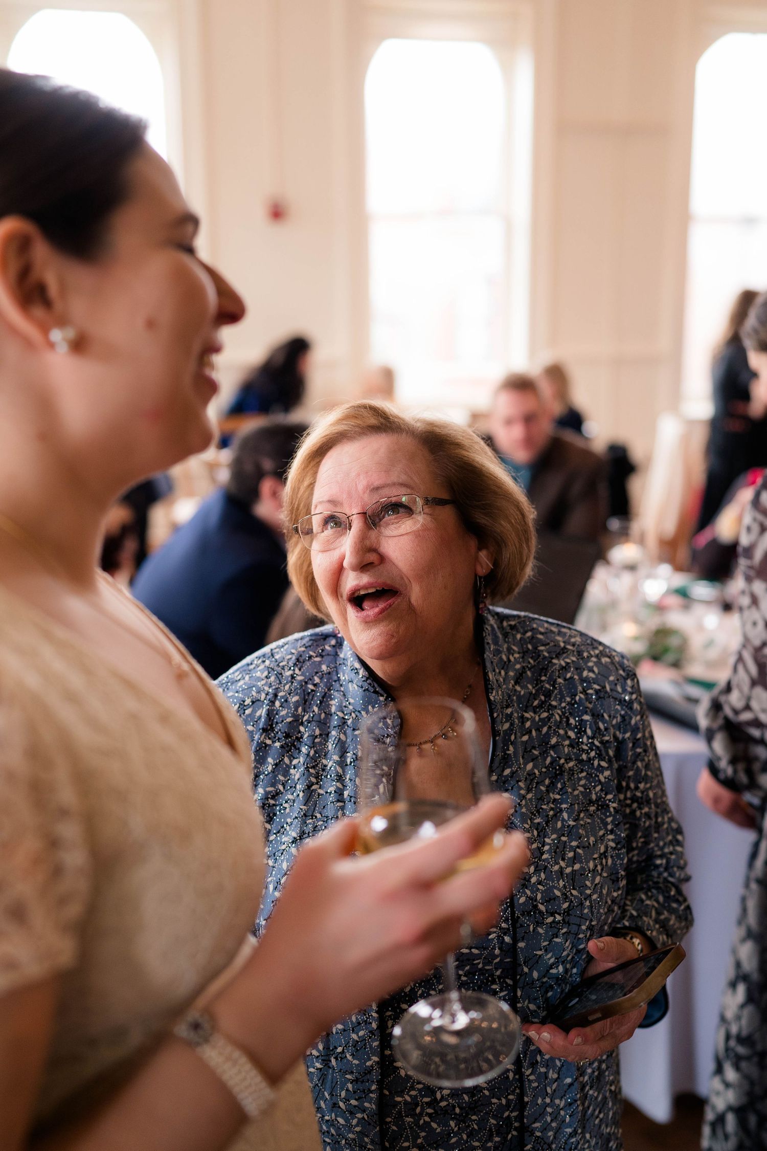 Two women sharing a lively conversation at a wedding reception, one holding a glass of white wine.
