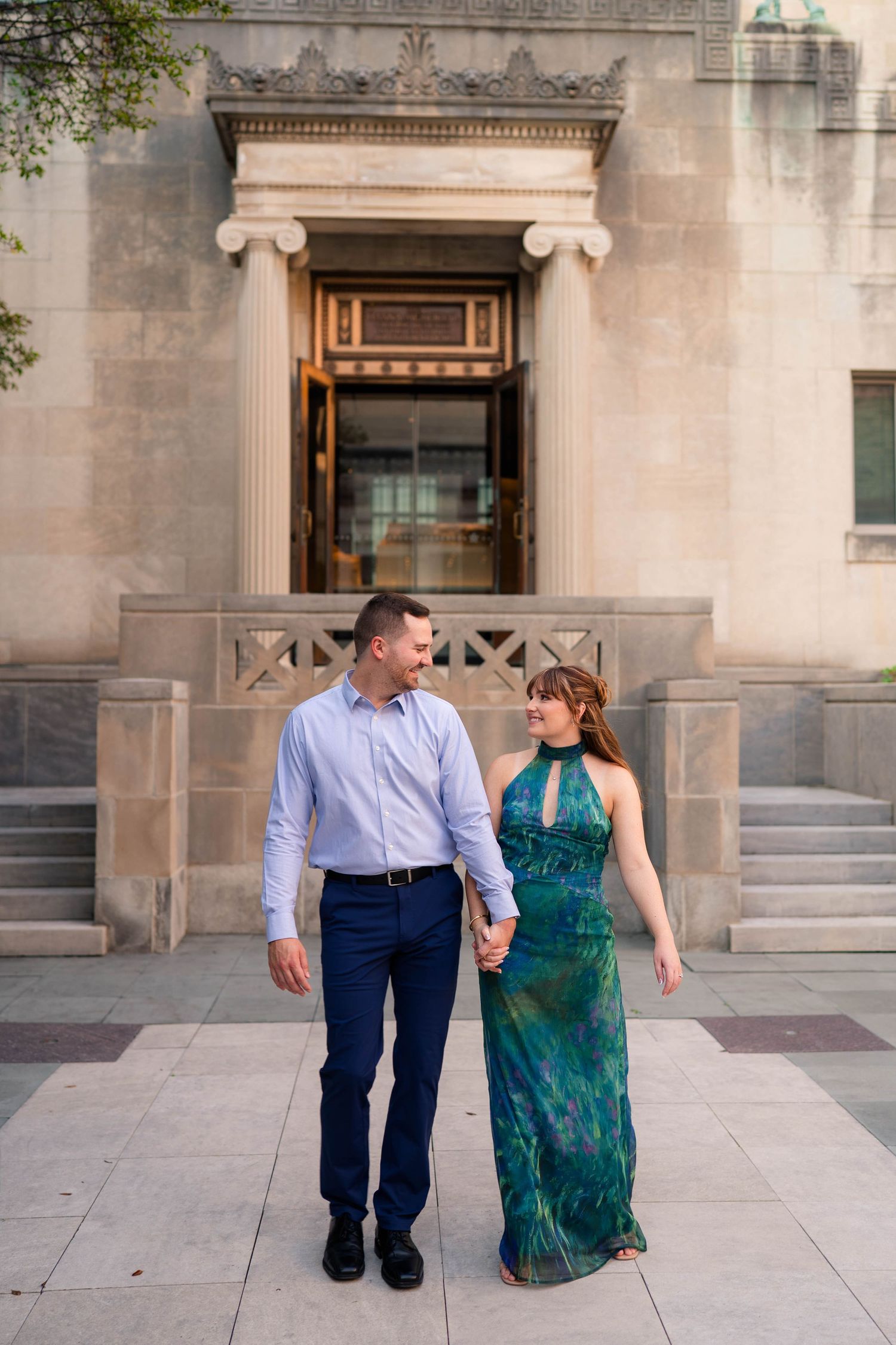 Couple holding hands walking in front of a neoclassical building, woman in green dress, man in blue shirt.