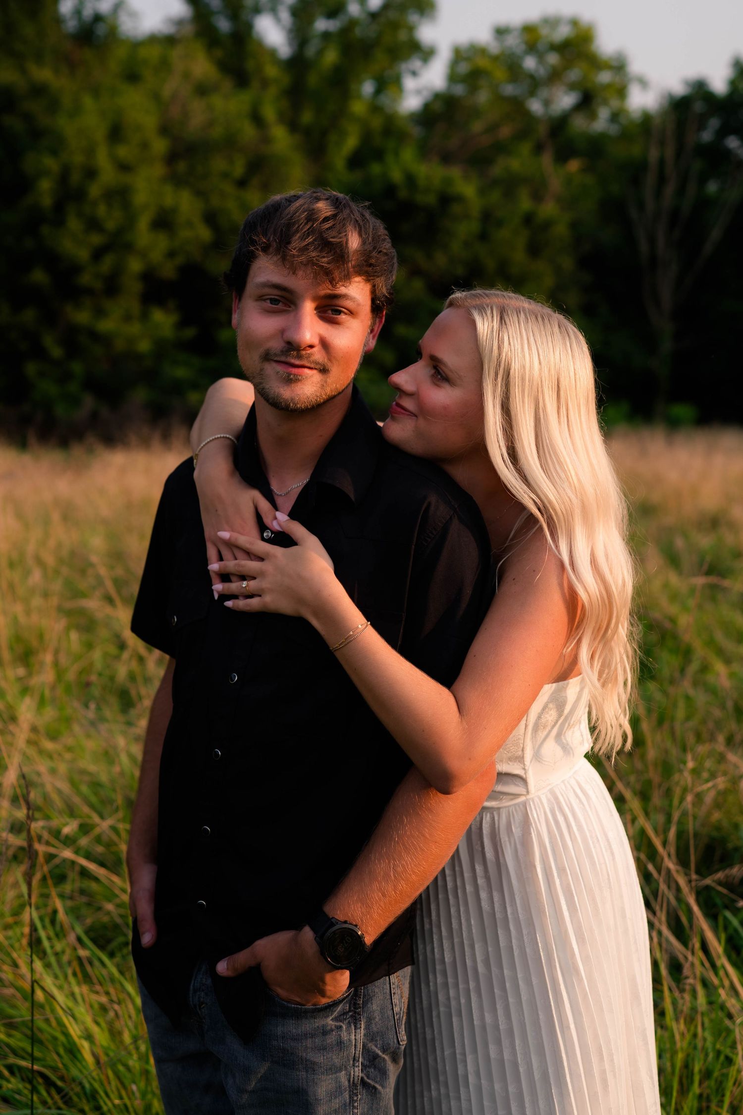 Couple embracing in golden field at sunset, woman in white dress hugging man in black shirt.