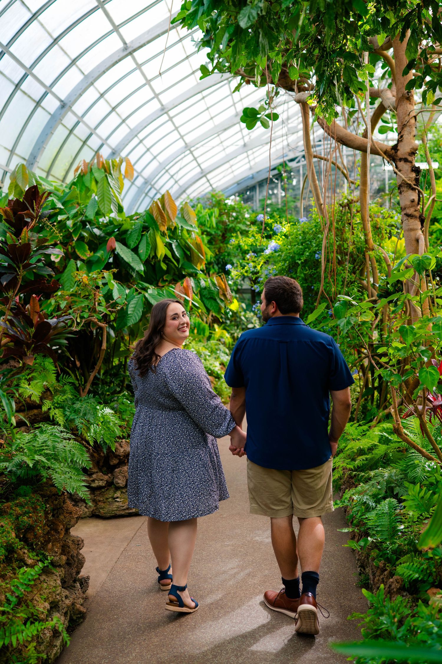 Couple holding hands walking through a lush tropical greenhouse conservatory pathway.