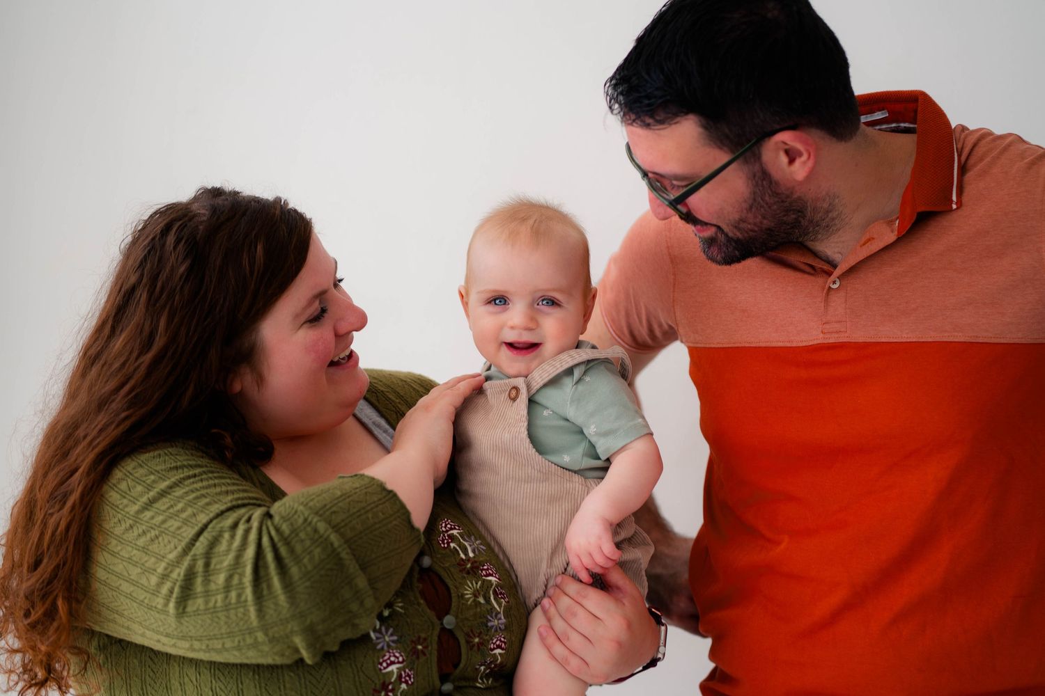 Happy family portrait with mother, father, and smiling baby in a bright white studio setting.