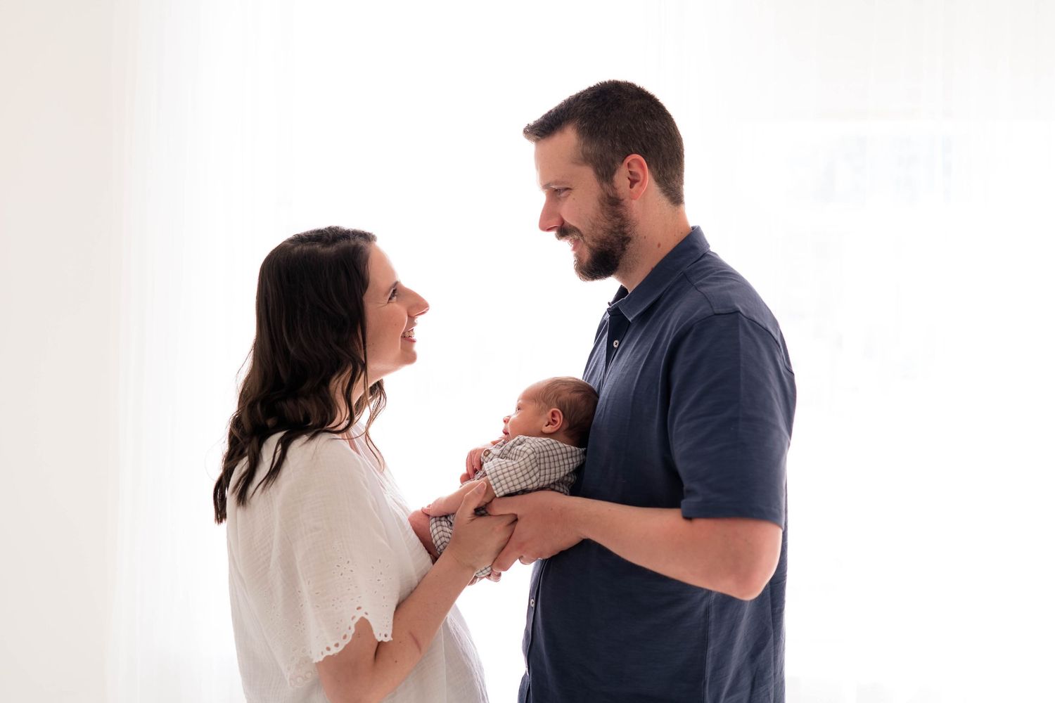 Happy new parents smiling at their newborn baby in a bright white studio setting.