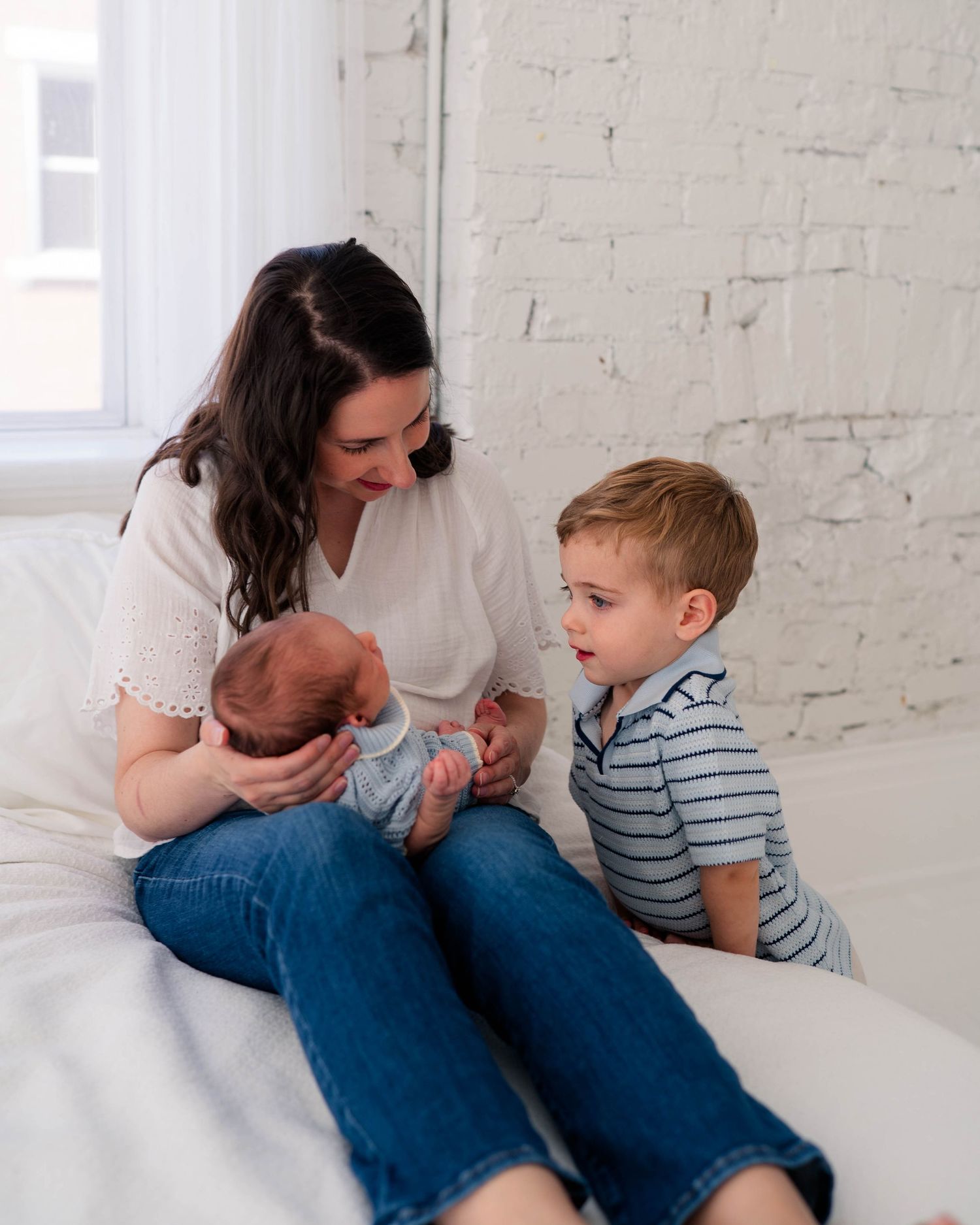 Mother sitting on white couch holding newborn while toddler boy looks on curiously.