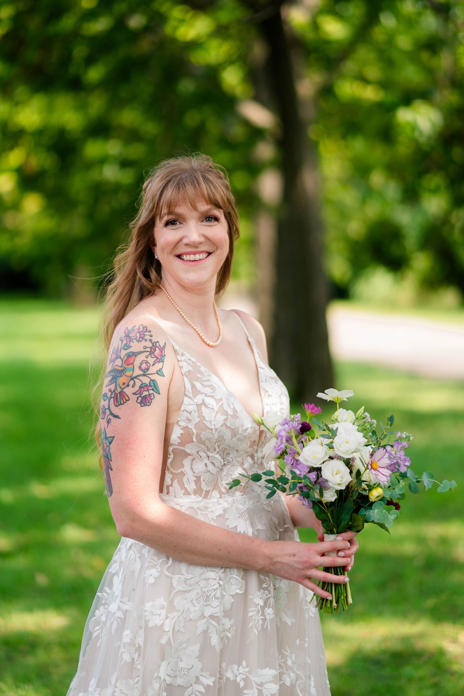 Smiling bride with floral arm tattoo in lace wedding gown holding colorful bouquet in park.
