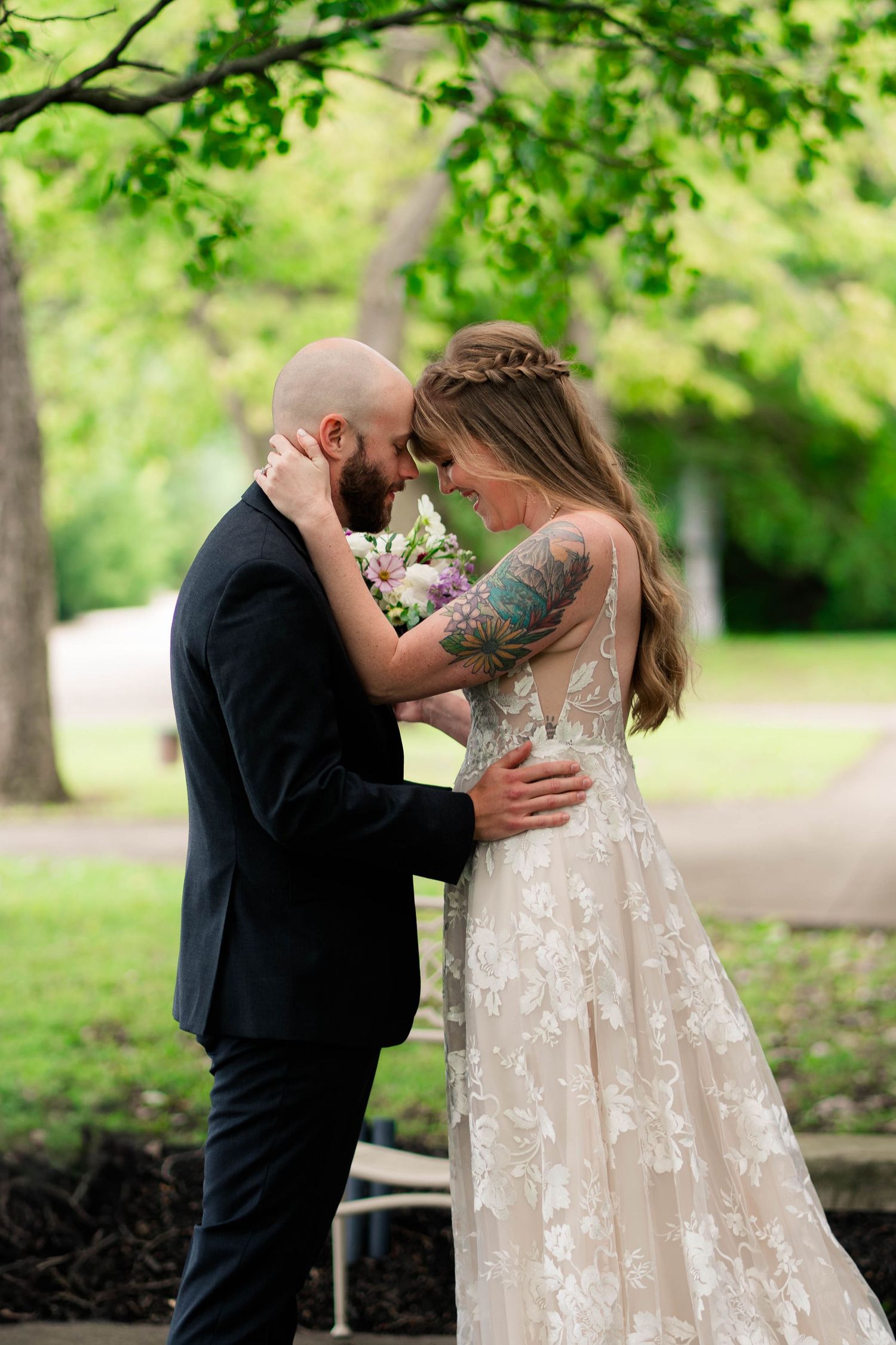A bride and groom share an intimate forehead touch moment surrounded by lush green trees outdoors.