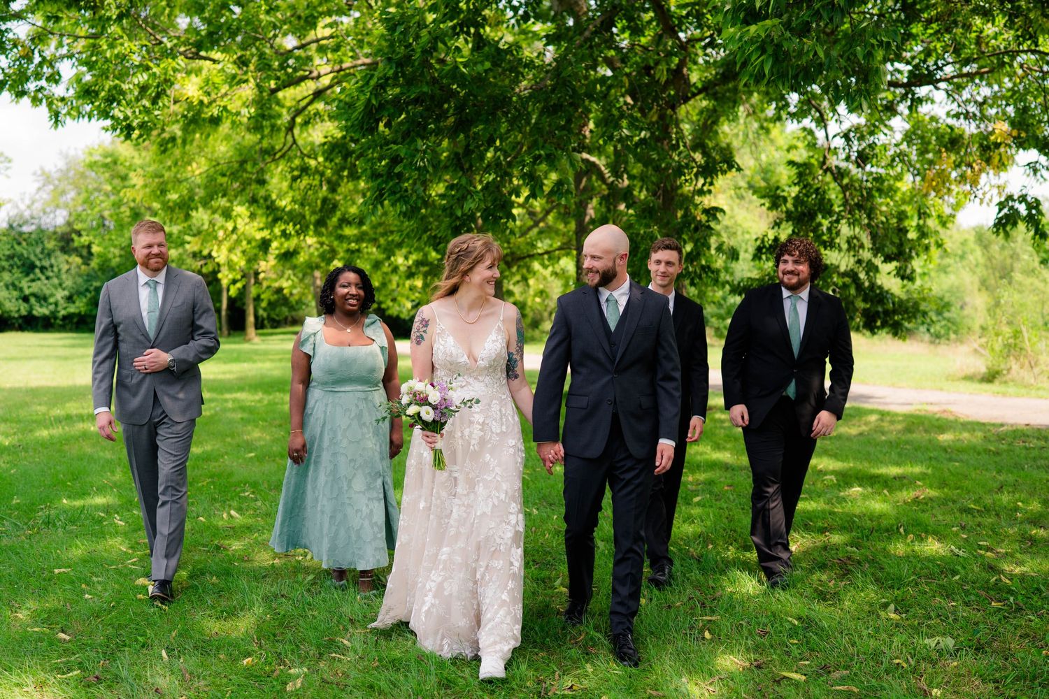 Wedding party walks together on green grass with bride in lace gown holding bouquet flanked by attendants.
