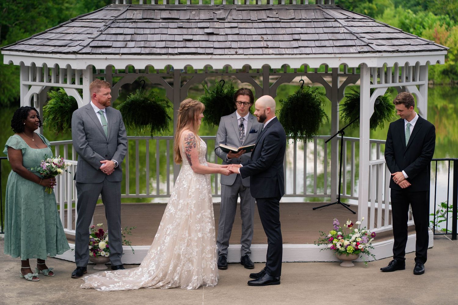 Bride and groom exchange vows at a white gazebo altar surrounded by wedding party members and lush greenery.