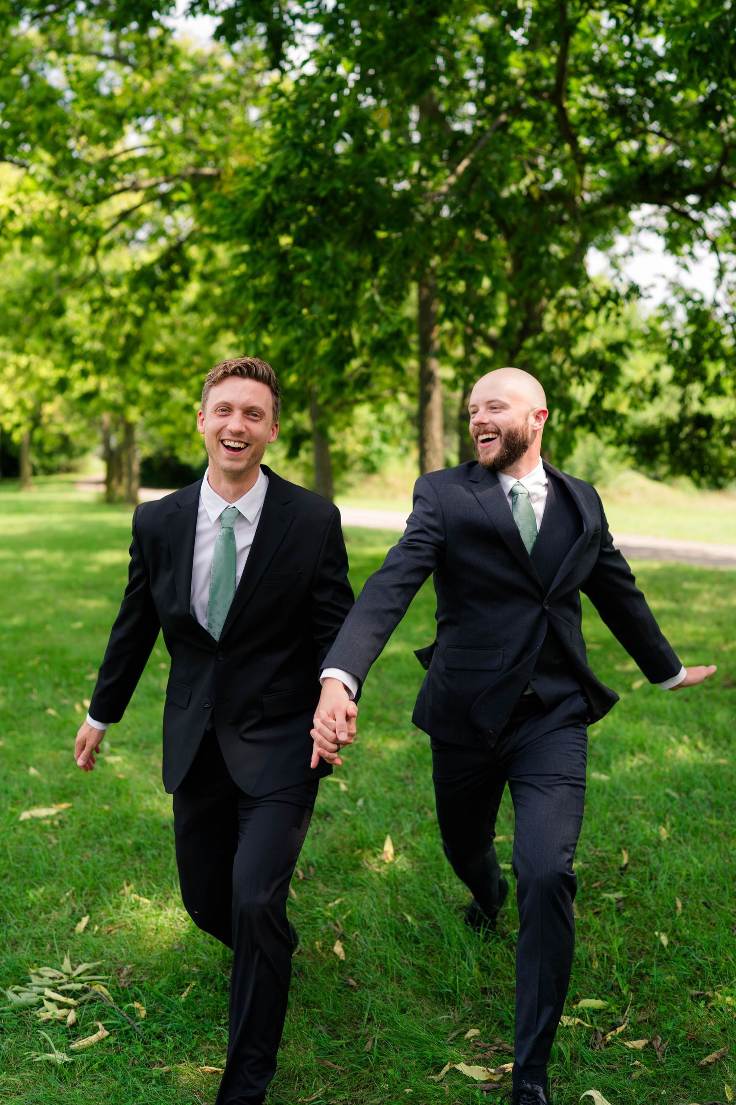 Two groomsmen in black suits with green ties laugh and hold hands while walking in park.