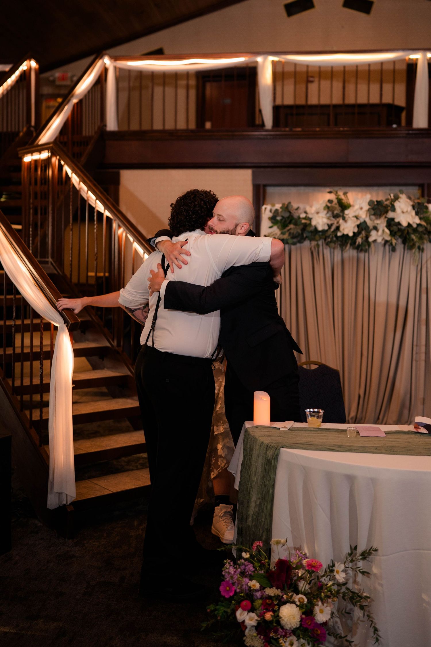 Two wedding guests share a warm embrace near a staircase at an elegant indoor wedding reception venue.