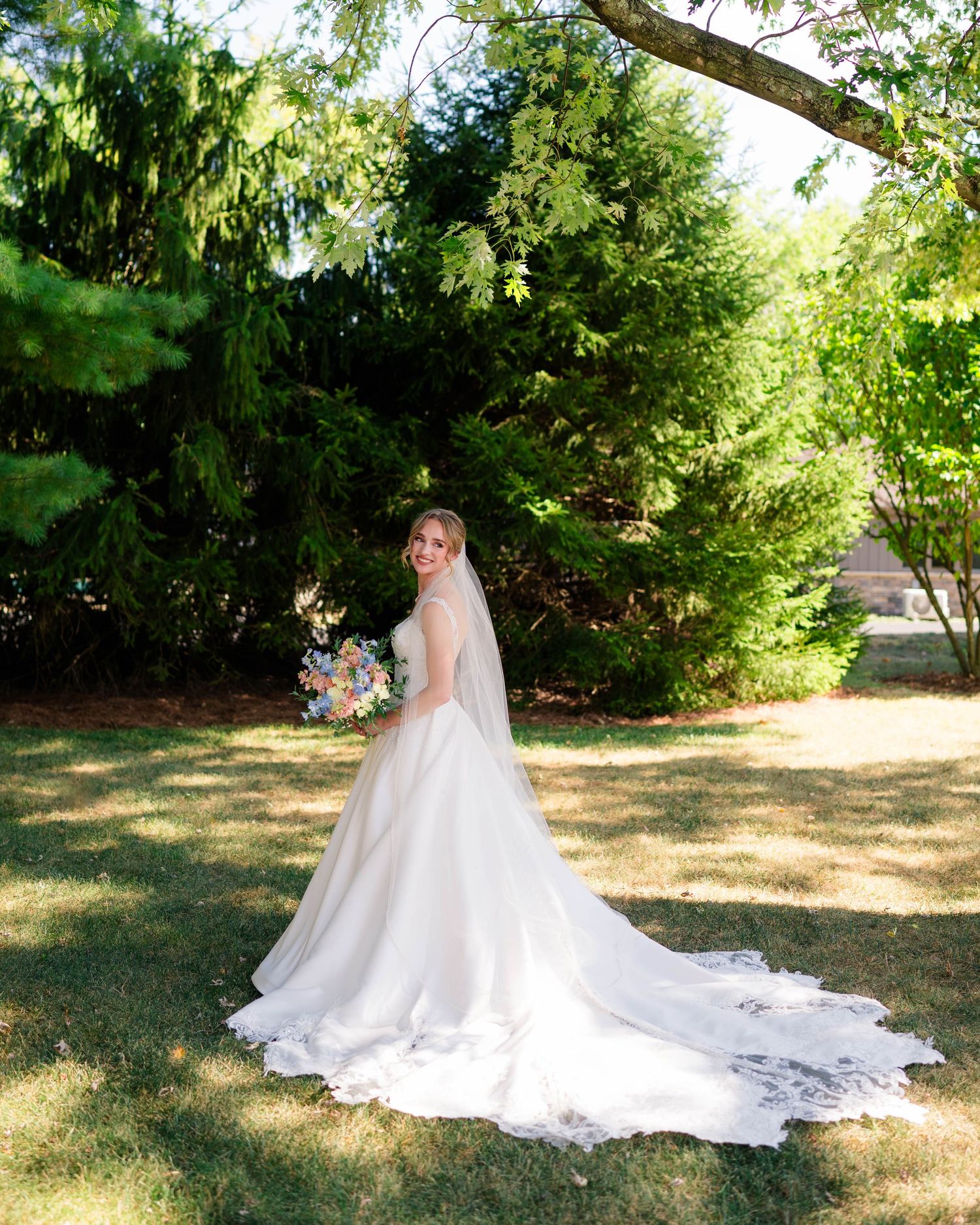 Bride in a long-train white lace wedding gown holding a colorful bouquet while posing in a lush garden.