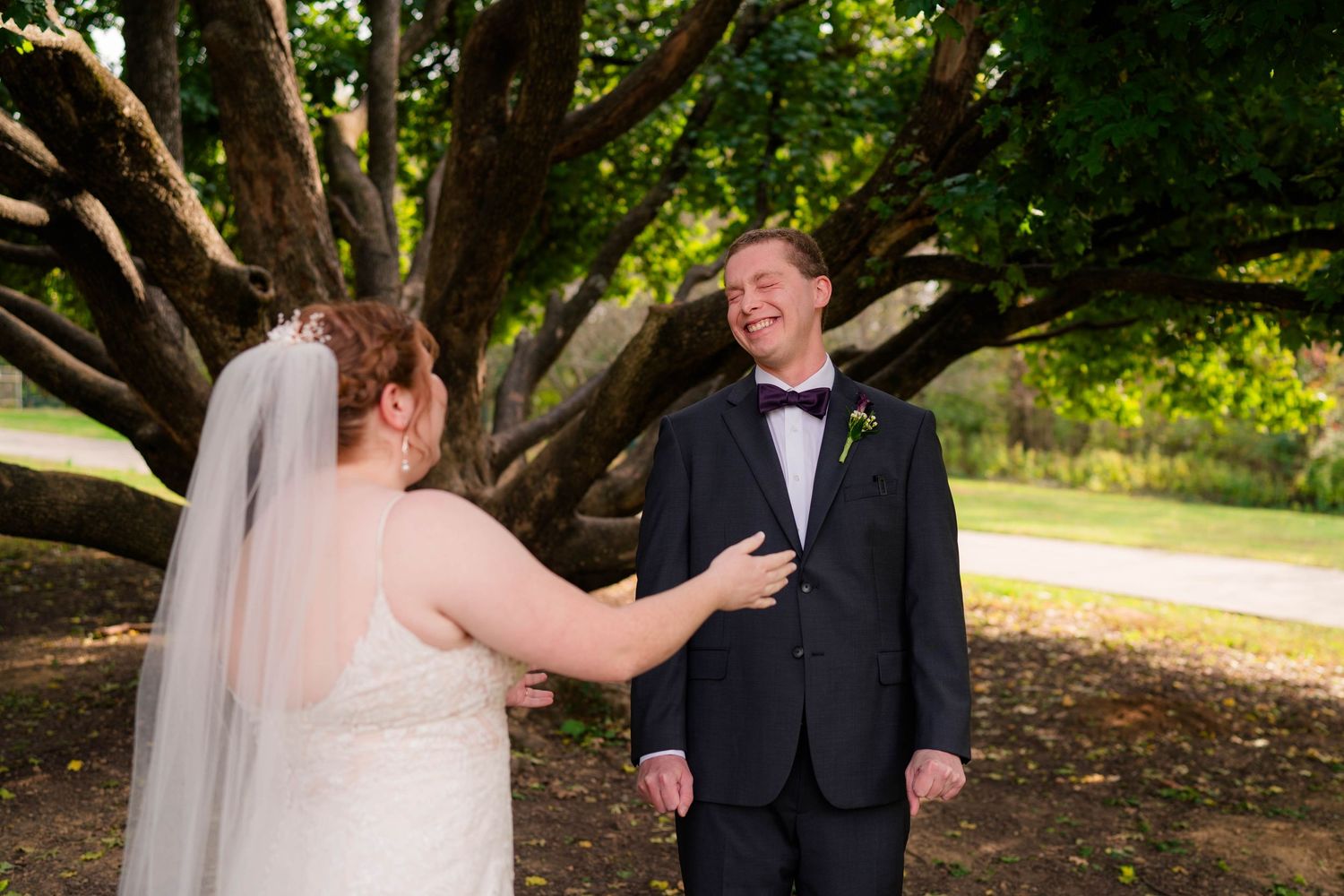 Groom smiles joyfully during first look moment with bride under large tree outdoors.