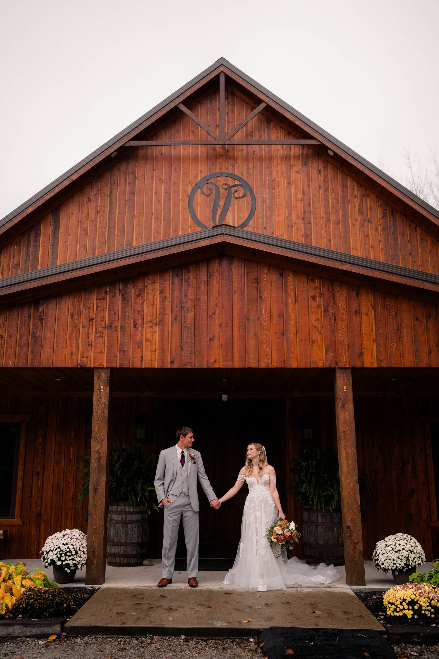 Bride and groom hold hands in front of a rustic wooden barn venue decorated with mums on their wedding day.