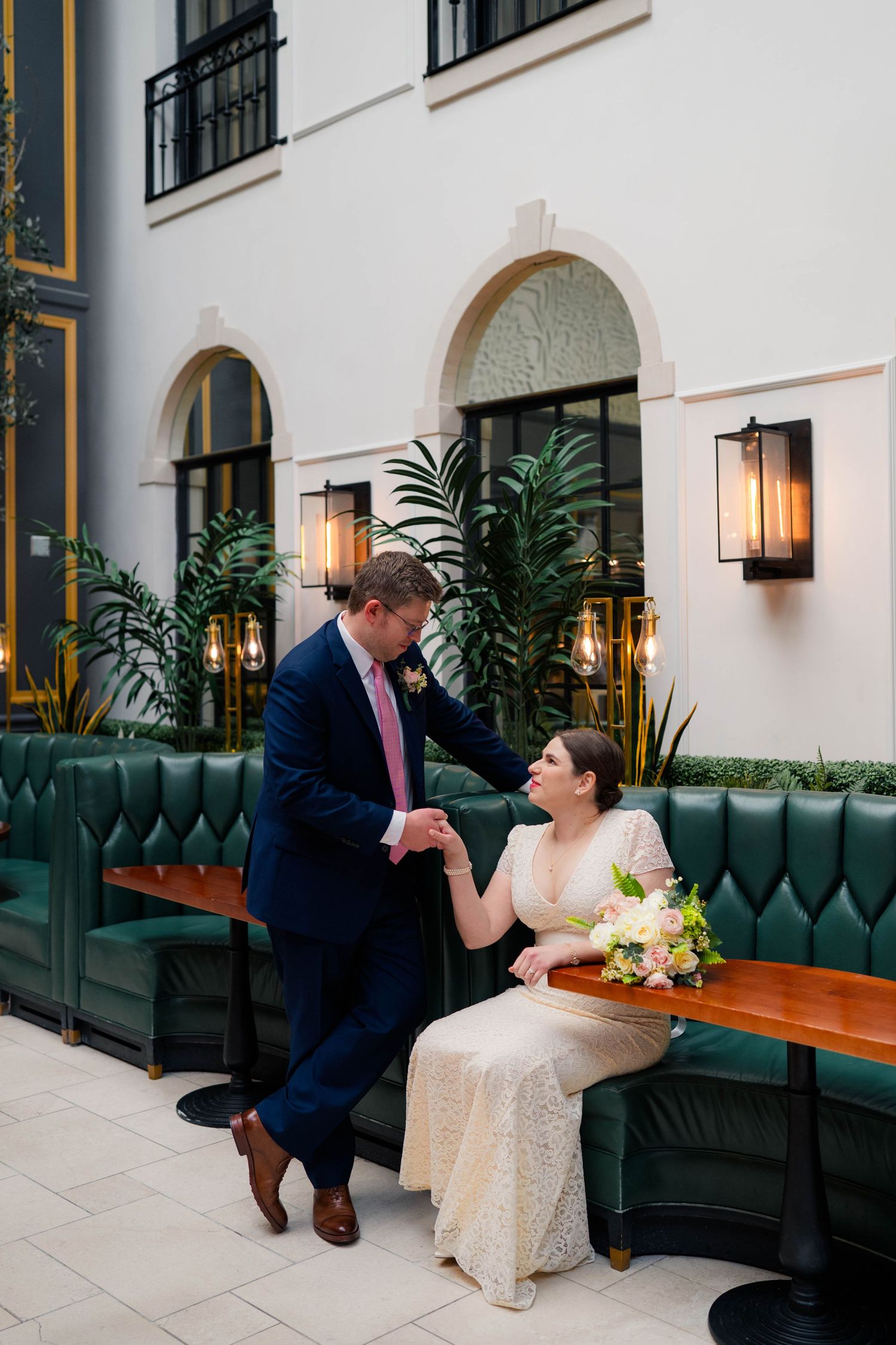 Groom in navy suit helps bride in lace dress rise from green tufted booth at wedding venue.