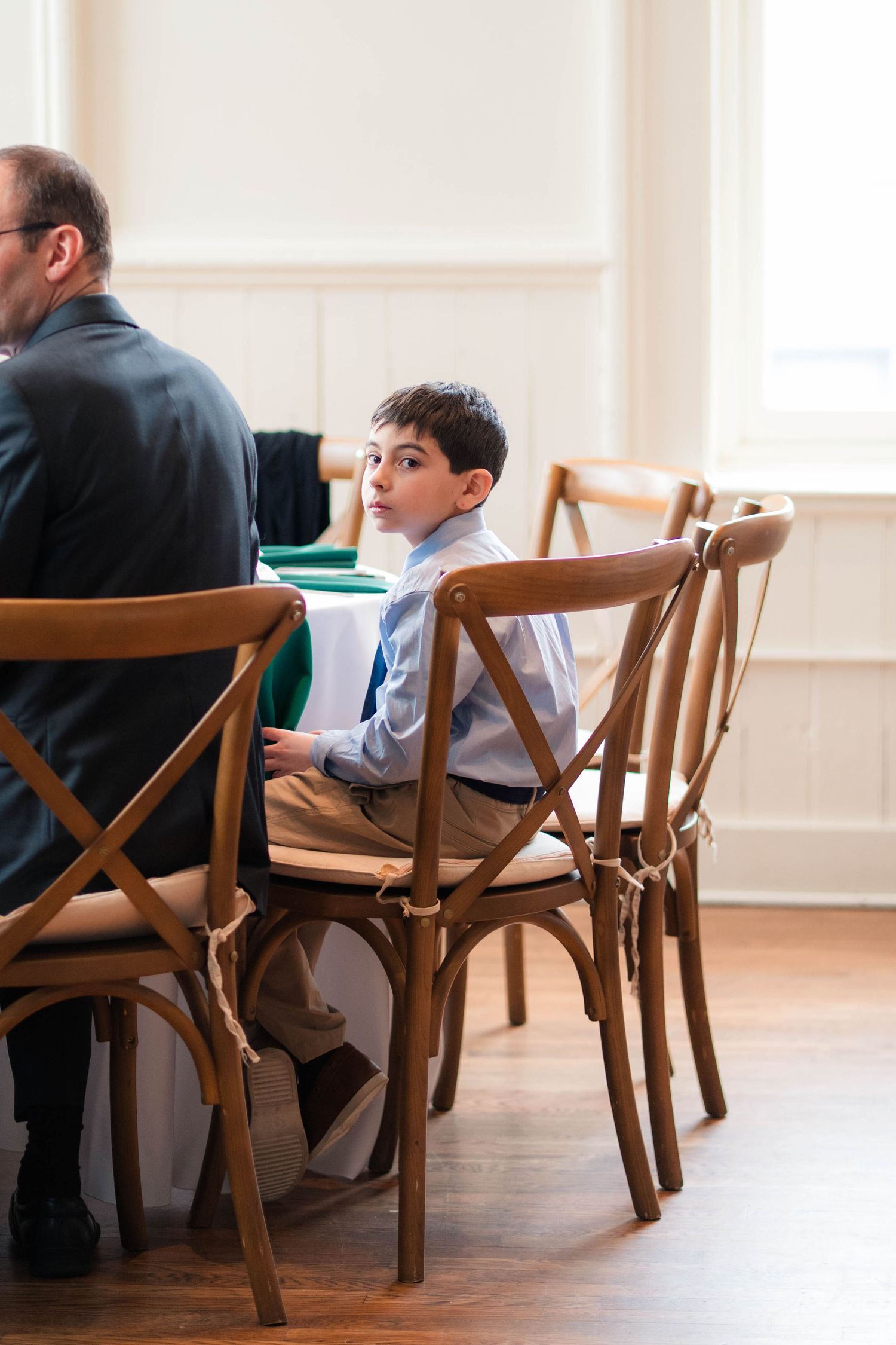 Young boy in dress shirt and tie sits attentively on a wooden cross-back chair at a formal event.