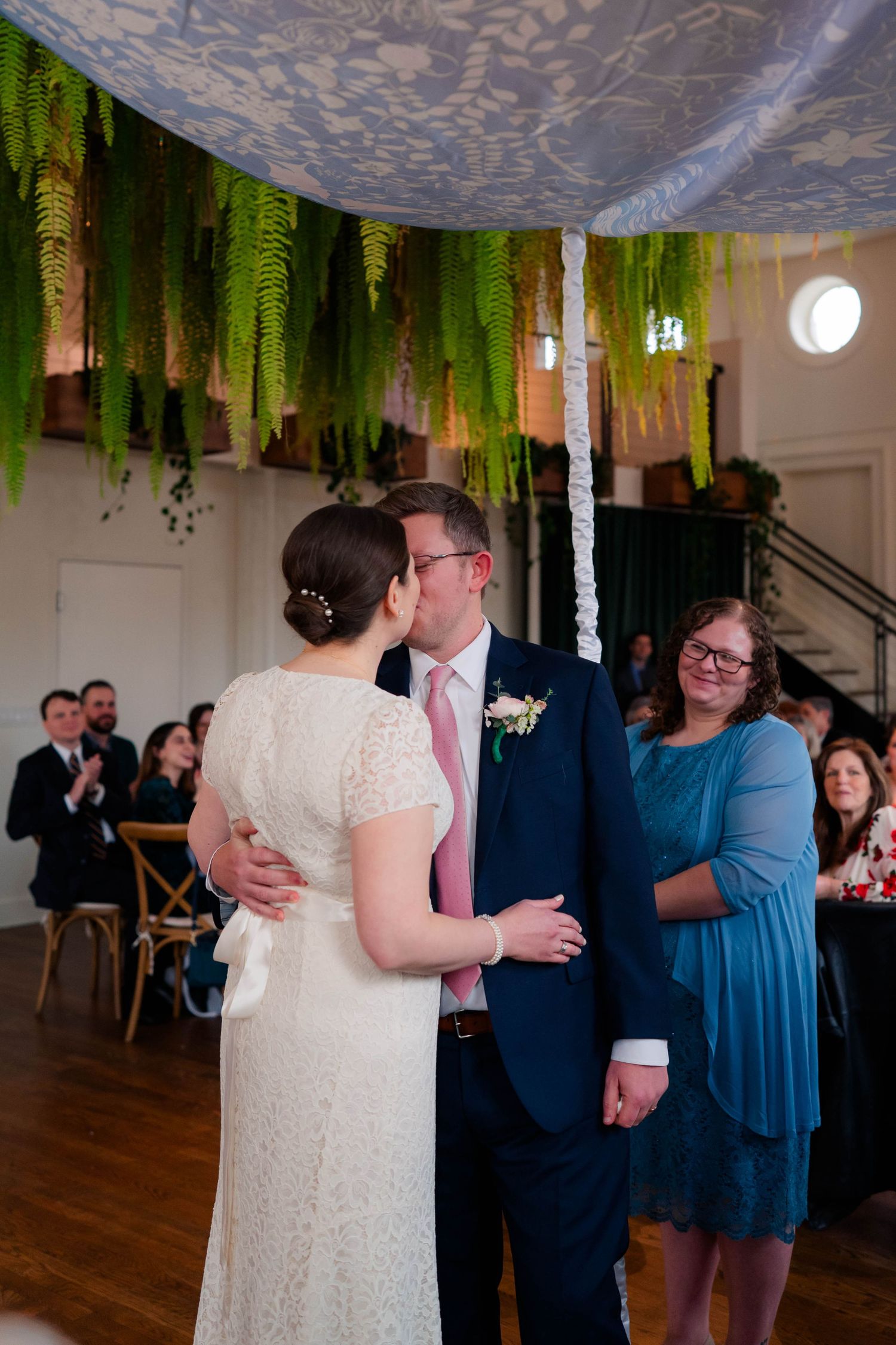 Newlyweds share a romantic kiss under a lush fern-adorned chuppah during indoor wedding ceremony.