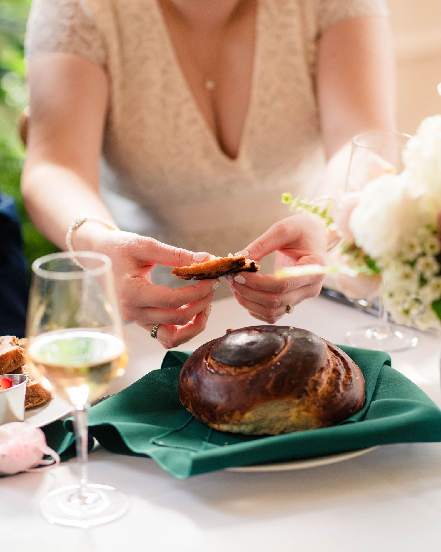 Bride in lace dress breaking traditional challah bread at wedding reception table with white wine and flowers.