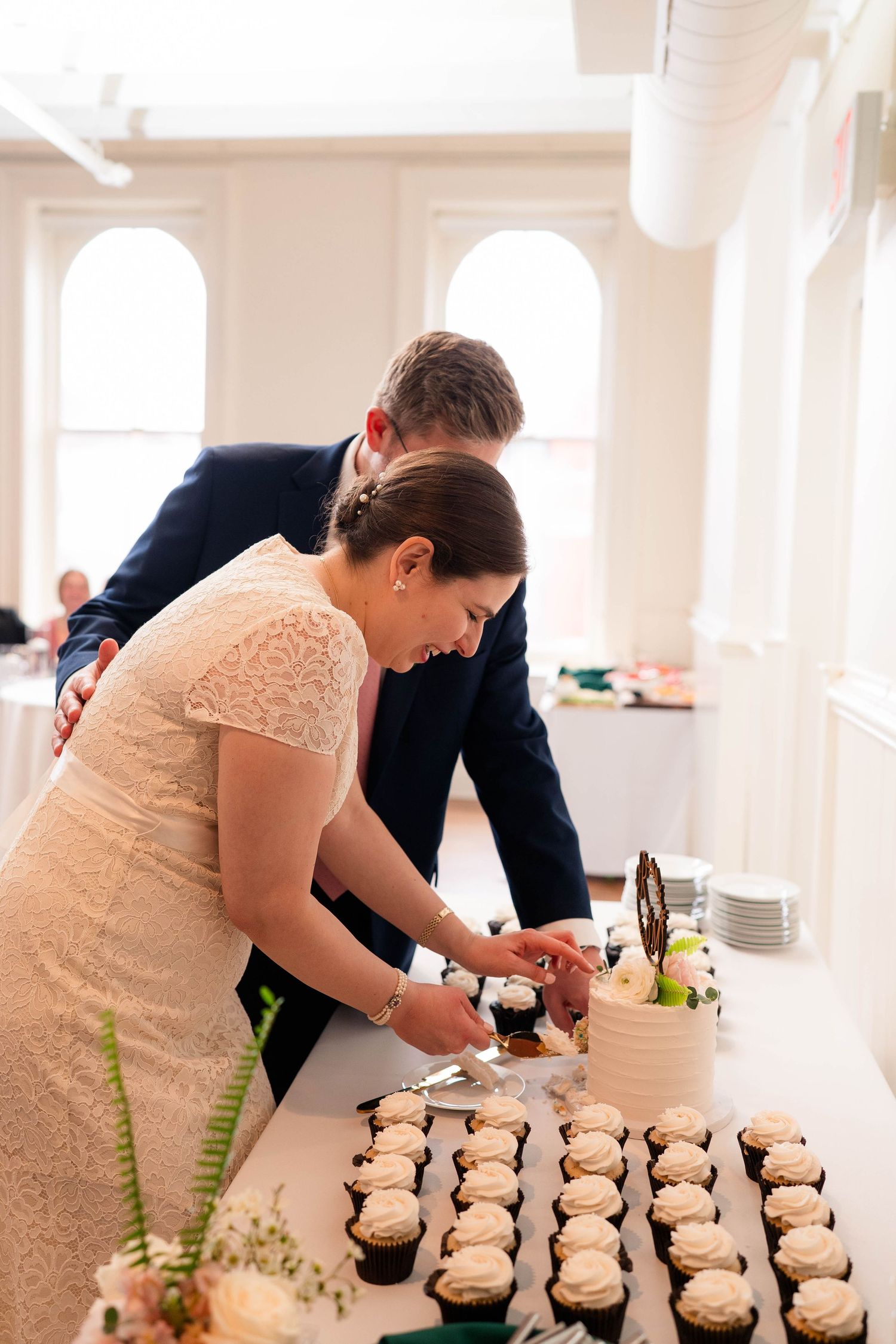 Bride and groom cutting their wedding cake together at reception table lined with white-frosted cupcakes.