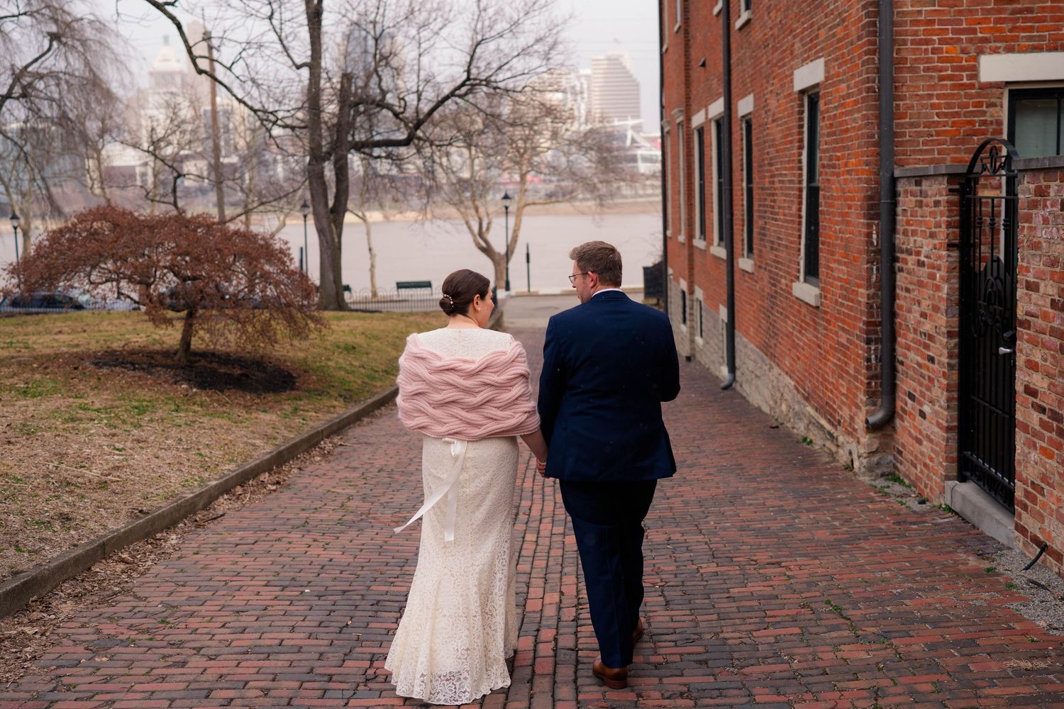 Bride and groom walk away hand-in-hand along a brick path beside a waterfront building on a misty day.
