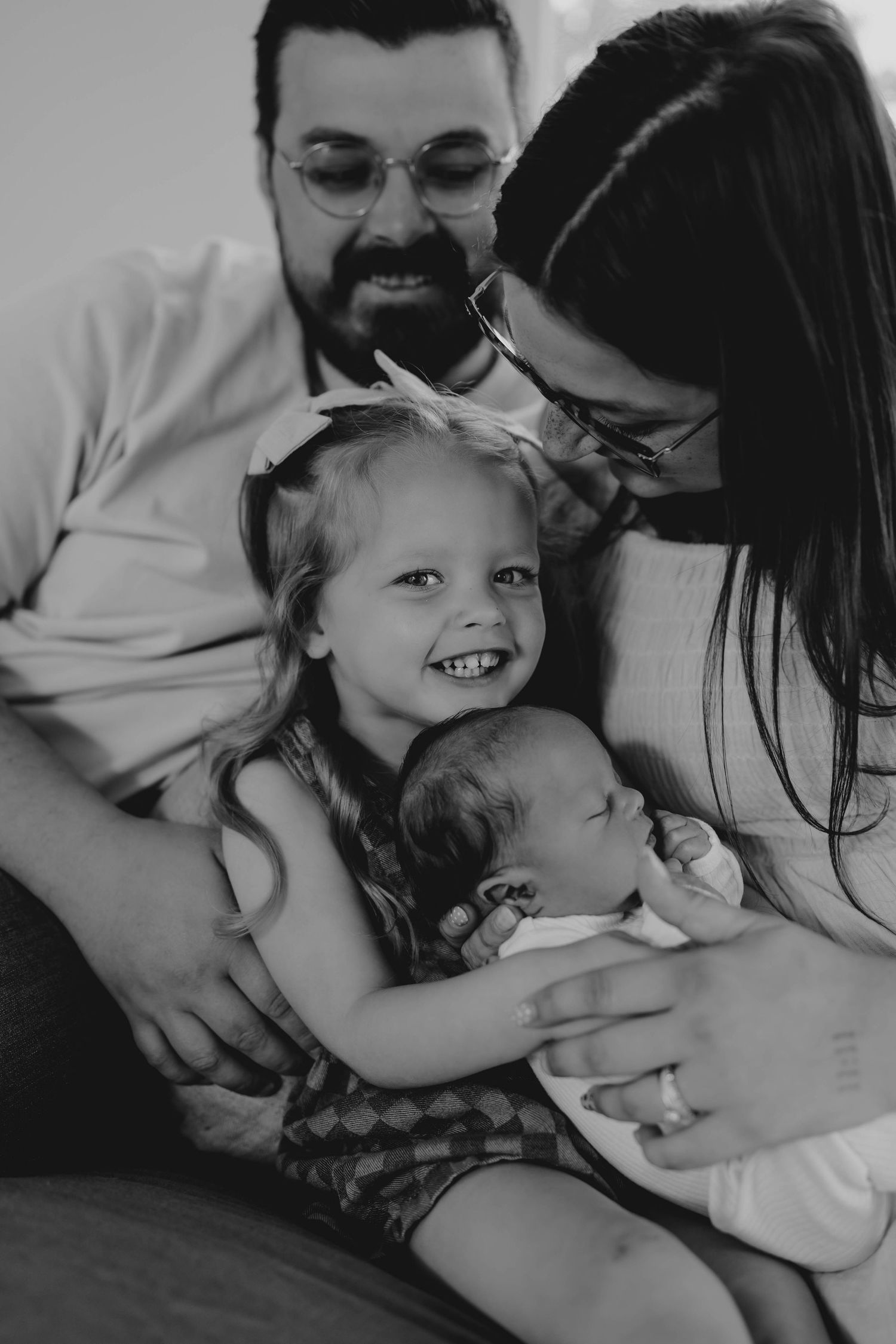 Black and white photo of smiling family with toddler girl holding newborn baby, parents leaning in lovingly.