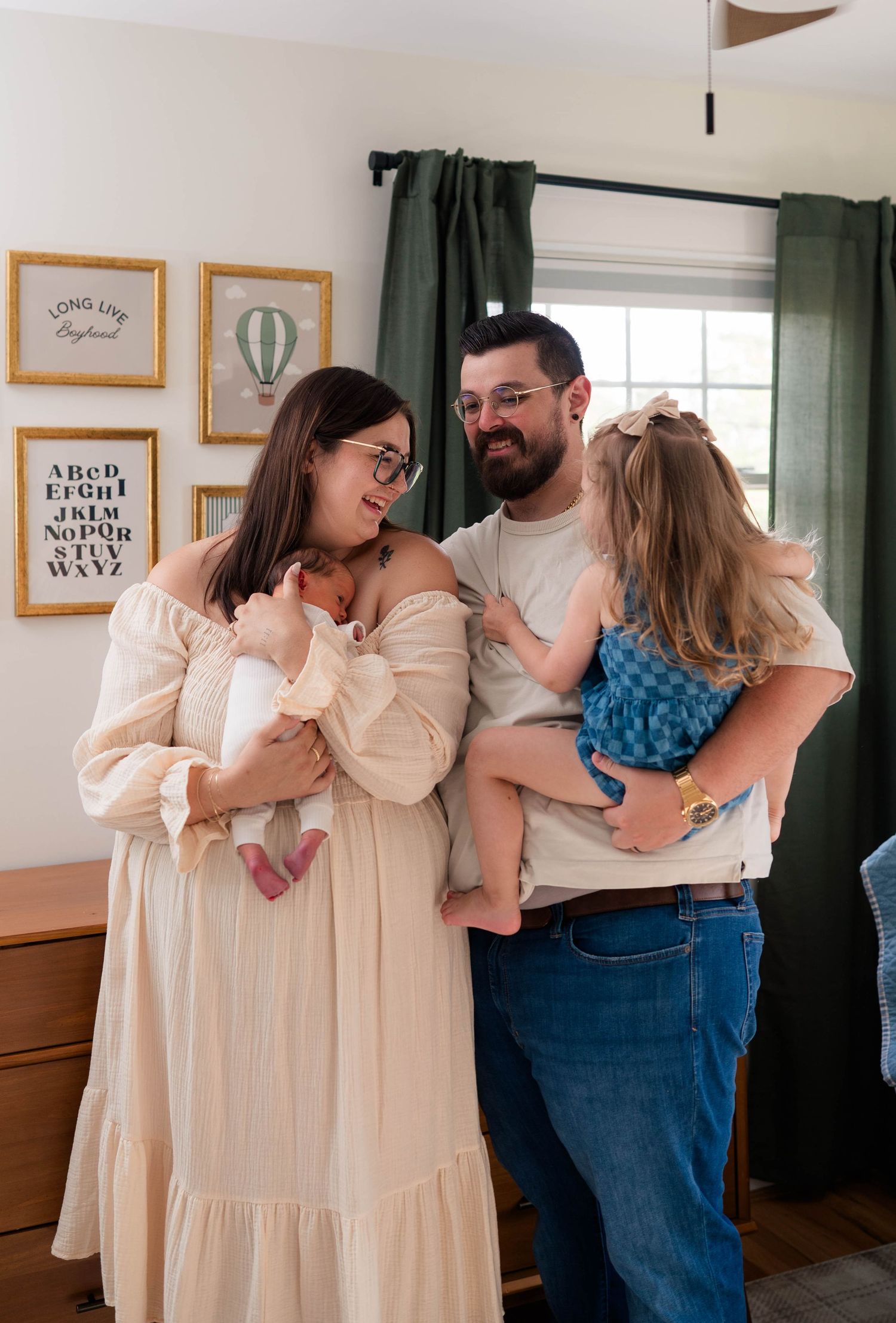 Joyful family of four laughs together in nursery, mom holding newborn and dad carrying toddler.