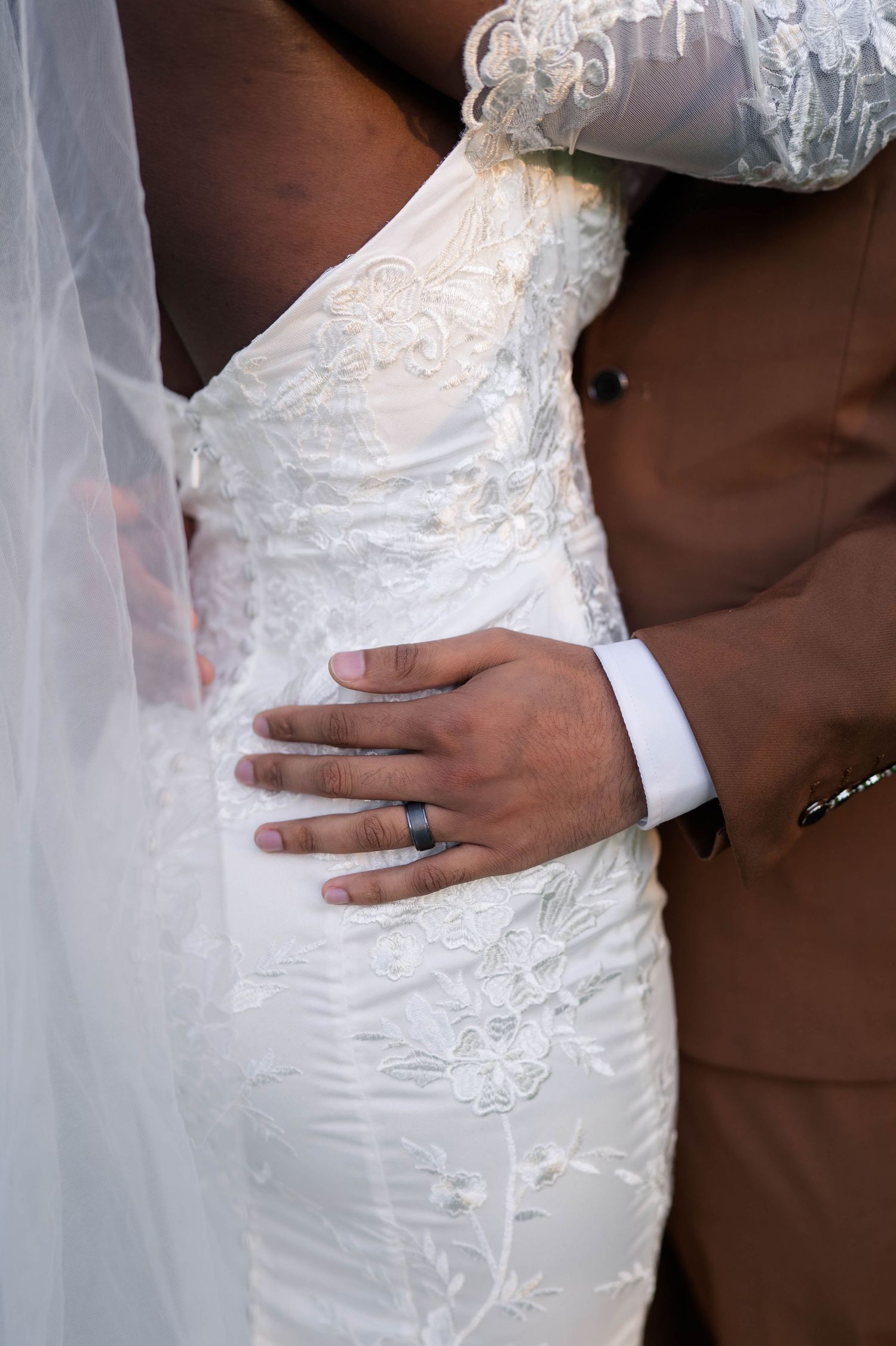 Groom in brown suit rests hand on brides white lace wedding dress, showing wedding band.