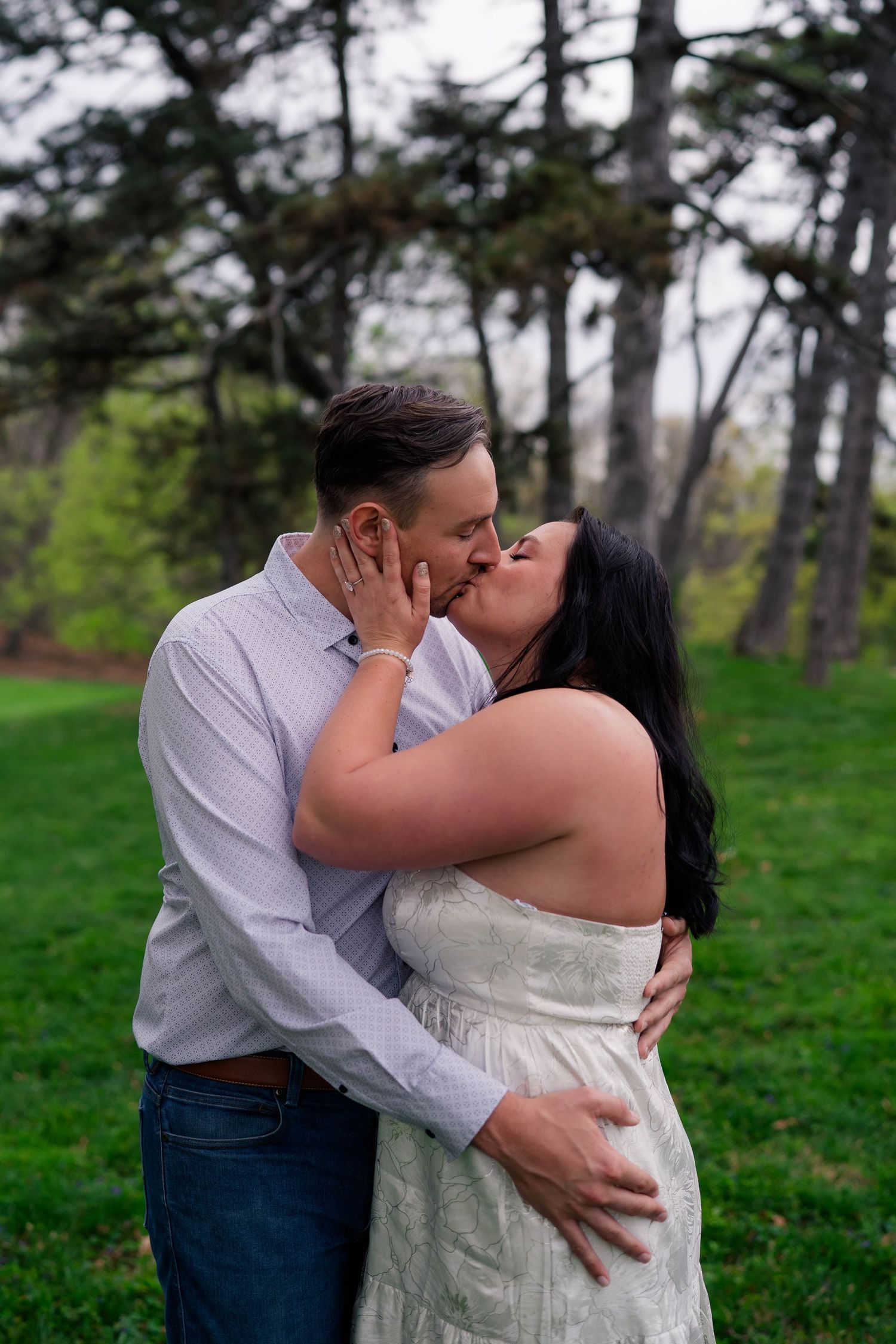A couple shares a romantic kiss outdoors on a green lawn surrounded by tall pine trees.