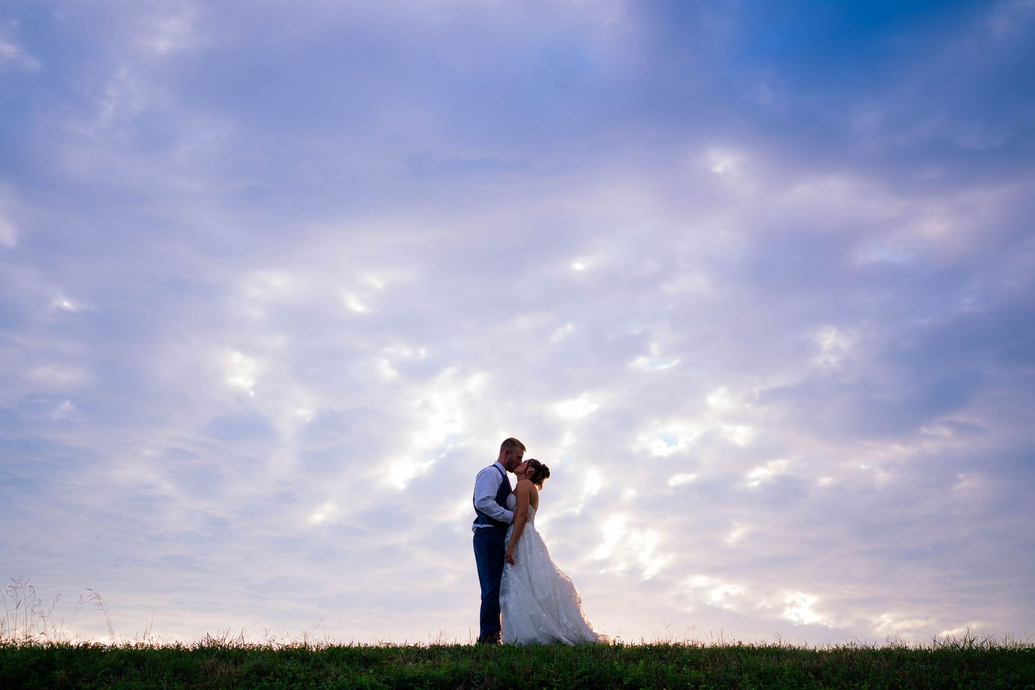 Silhouette of bride and groom kissing on a grassy hill under a dramatic purple cloudy sky at dusk.