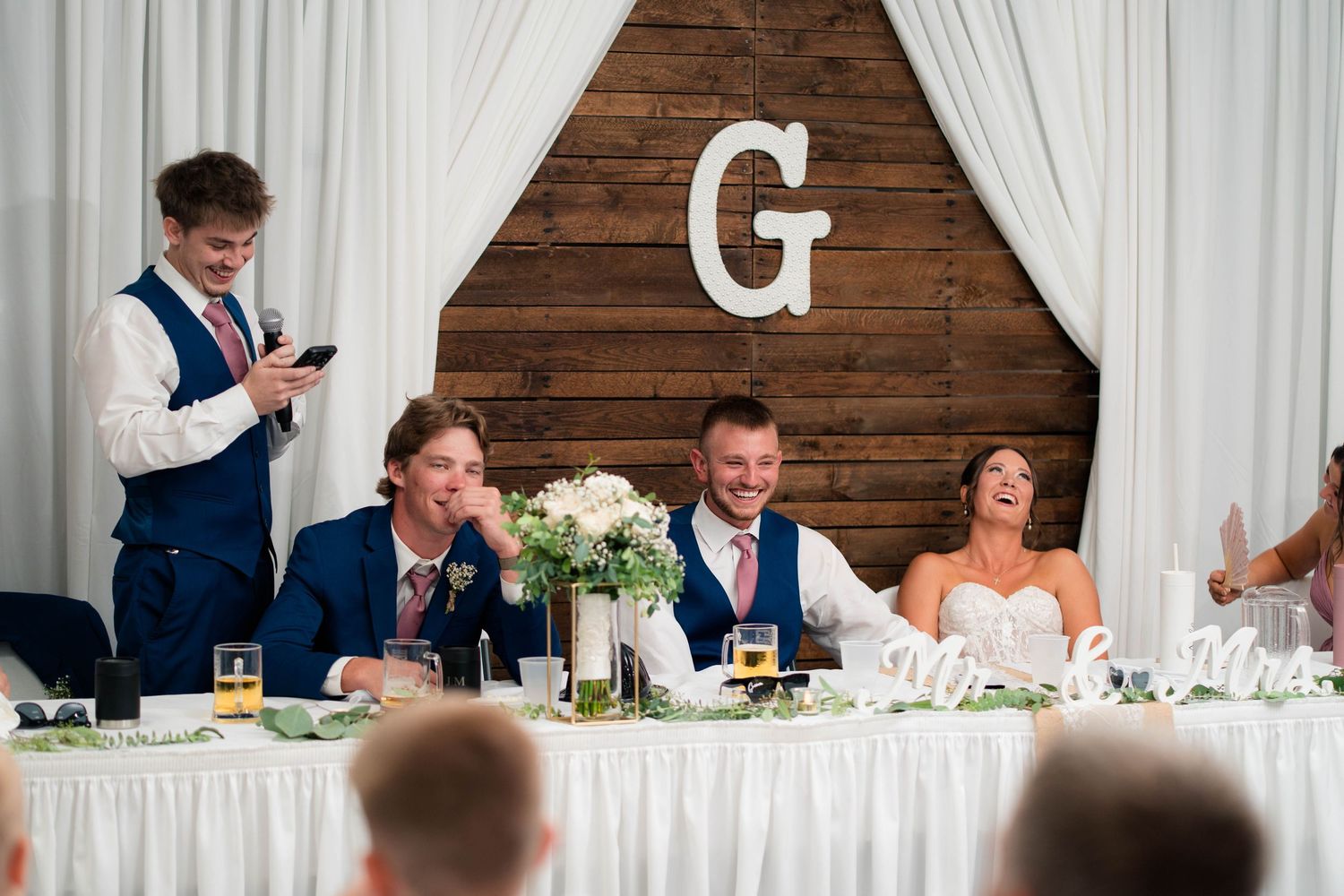 Groomsman reads phone while wedding party smiles at head table beneath wooden monogram G backdrop at reception.