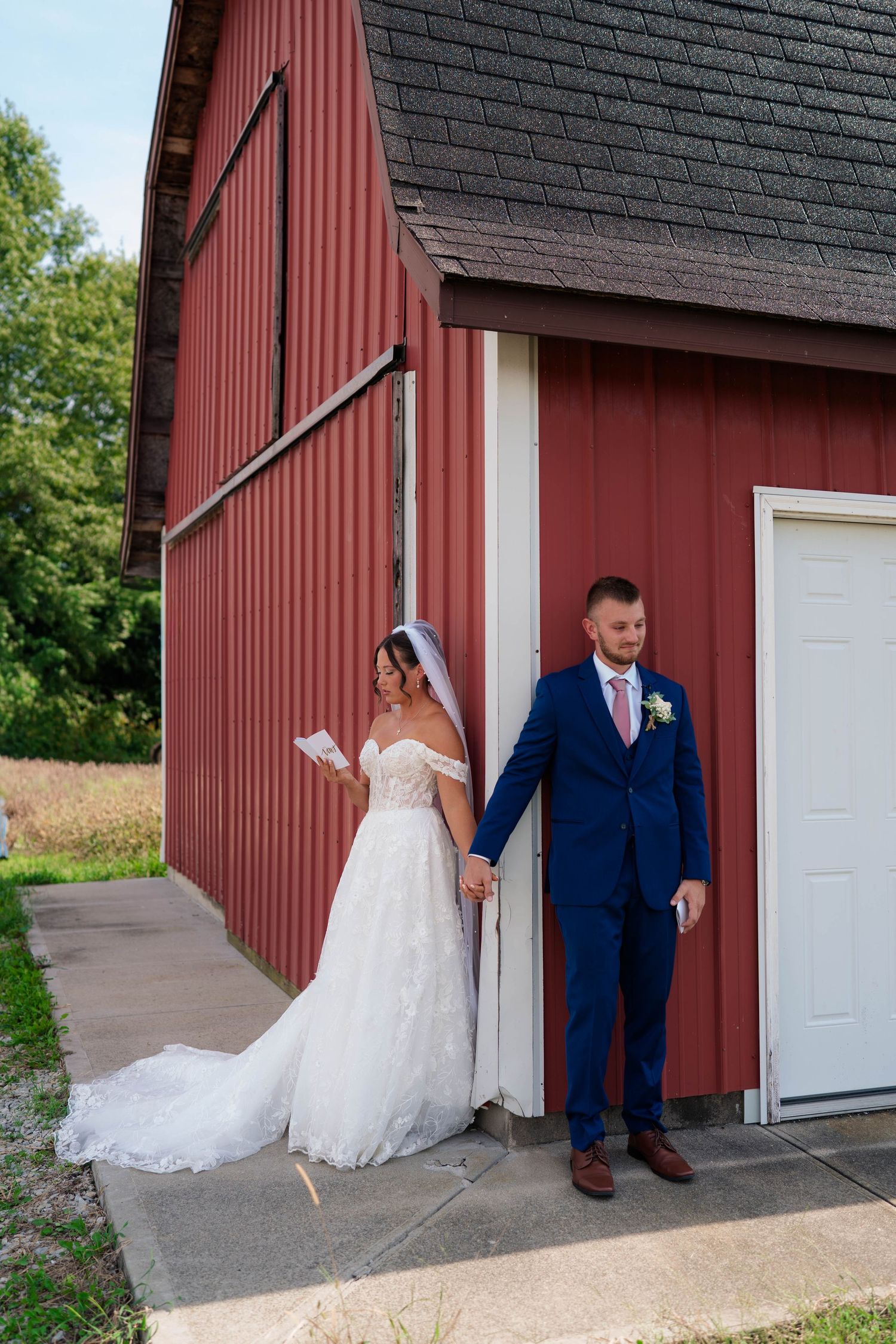 Bride reads vows while holding groom's hand around red barn corner on wedding day.