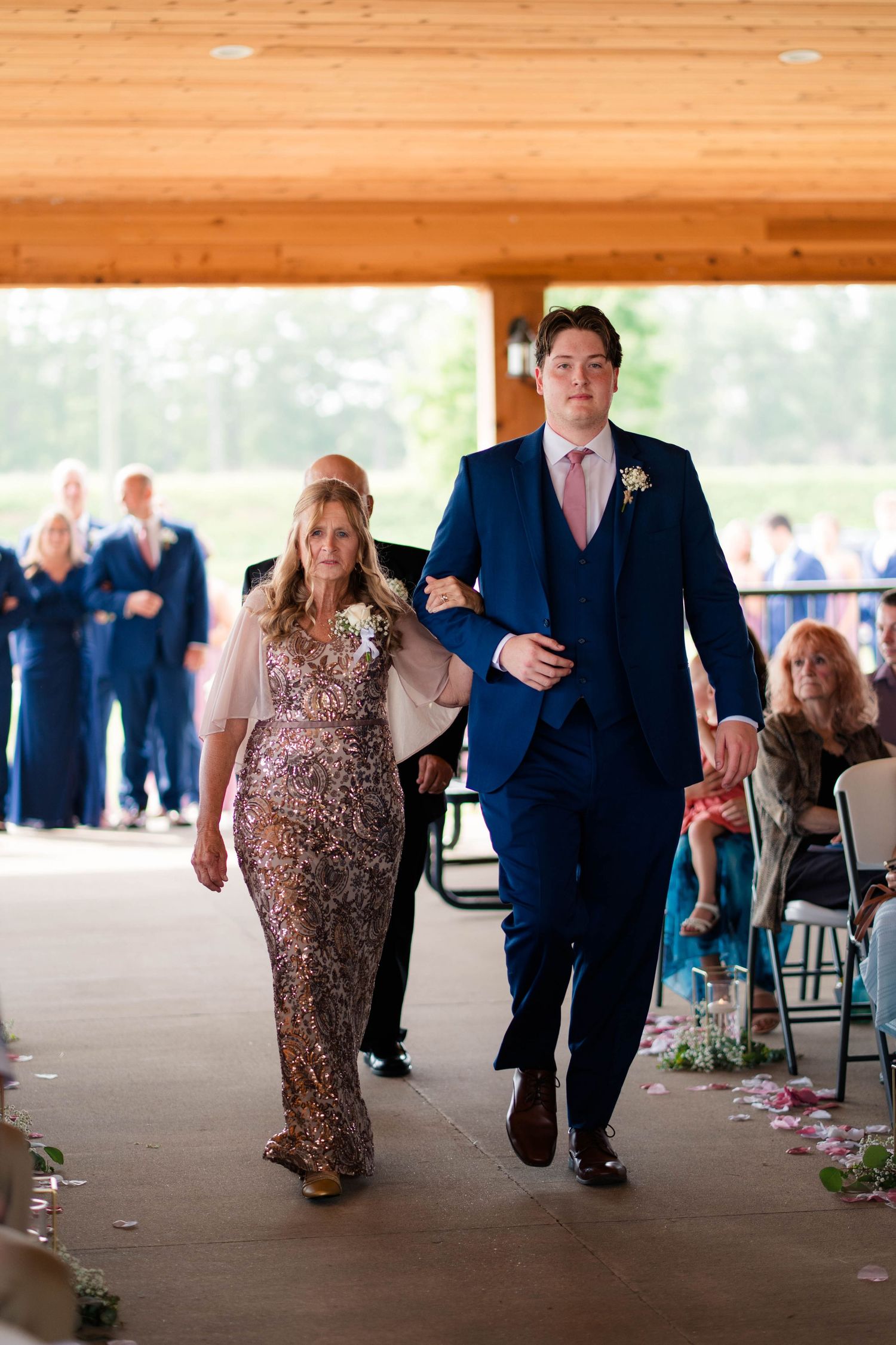 Groom in navy suit escorts elegantly dressed woman down wedding ceremony aisle under wooden pavilion.