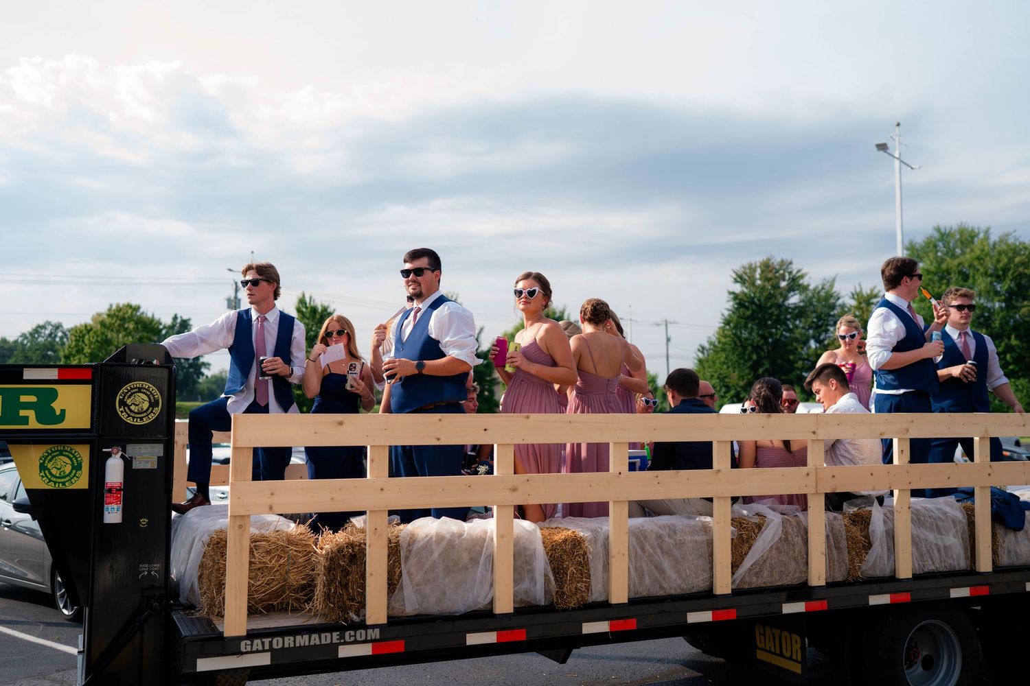 Wedding party rides on a flatbed truck with hay bales during a fun outdoor celebration.