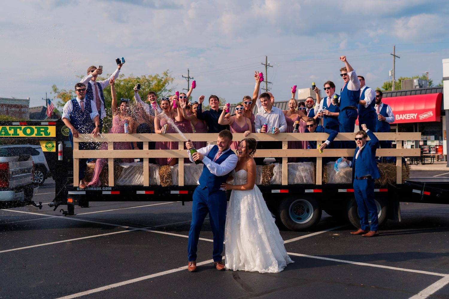 Bride and groom kiss in parking lot as joyful wedding party cheers from decorated flatbed truck.